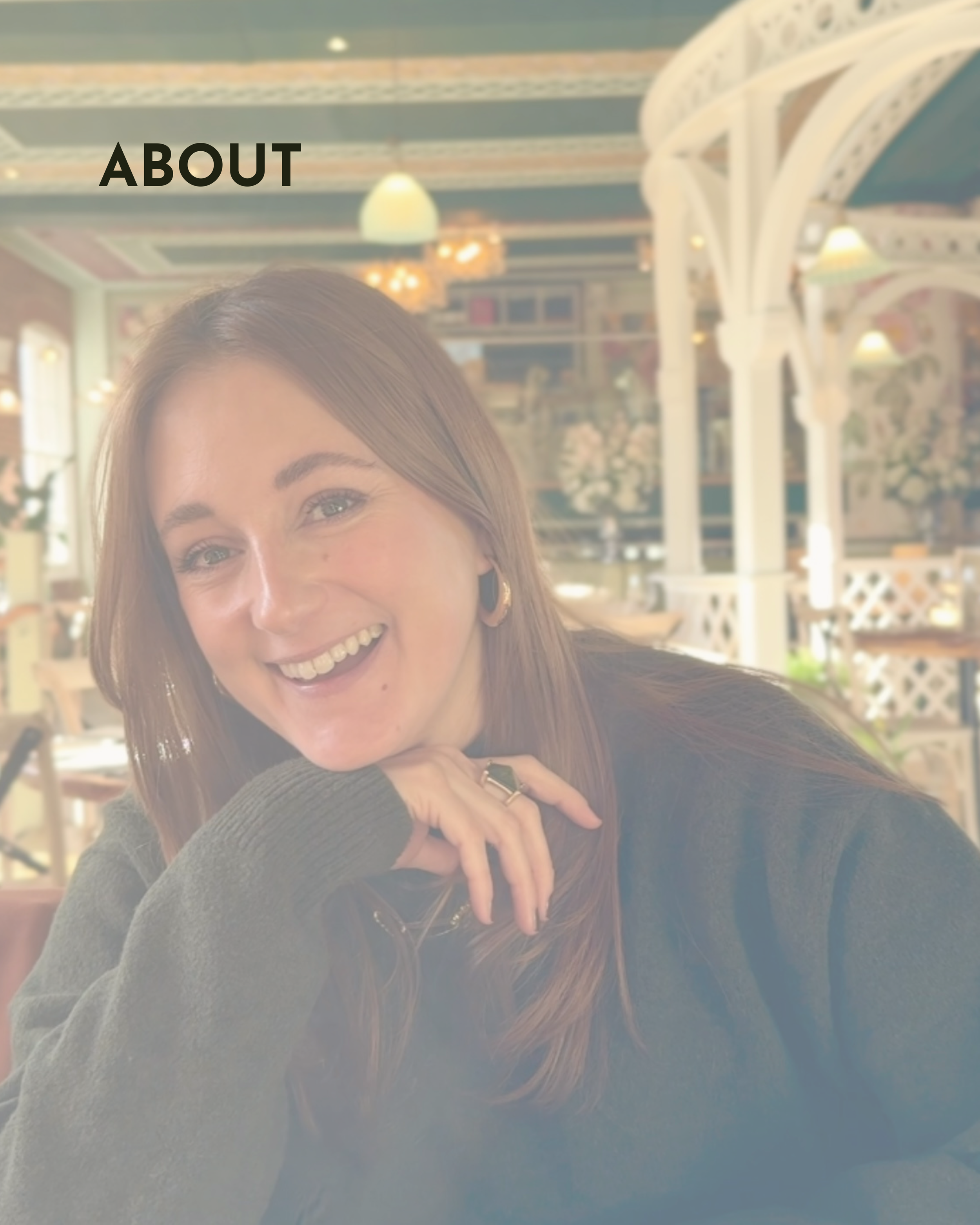 A brunette sat in a restaurant looking at the camera laughing and smiling