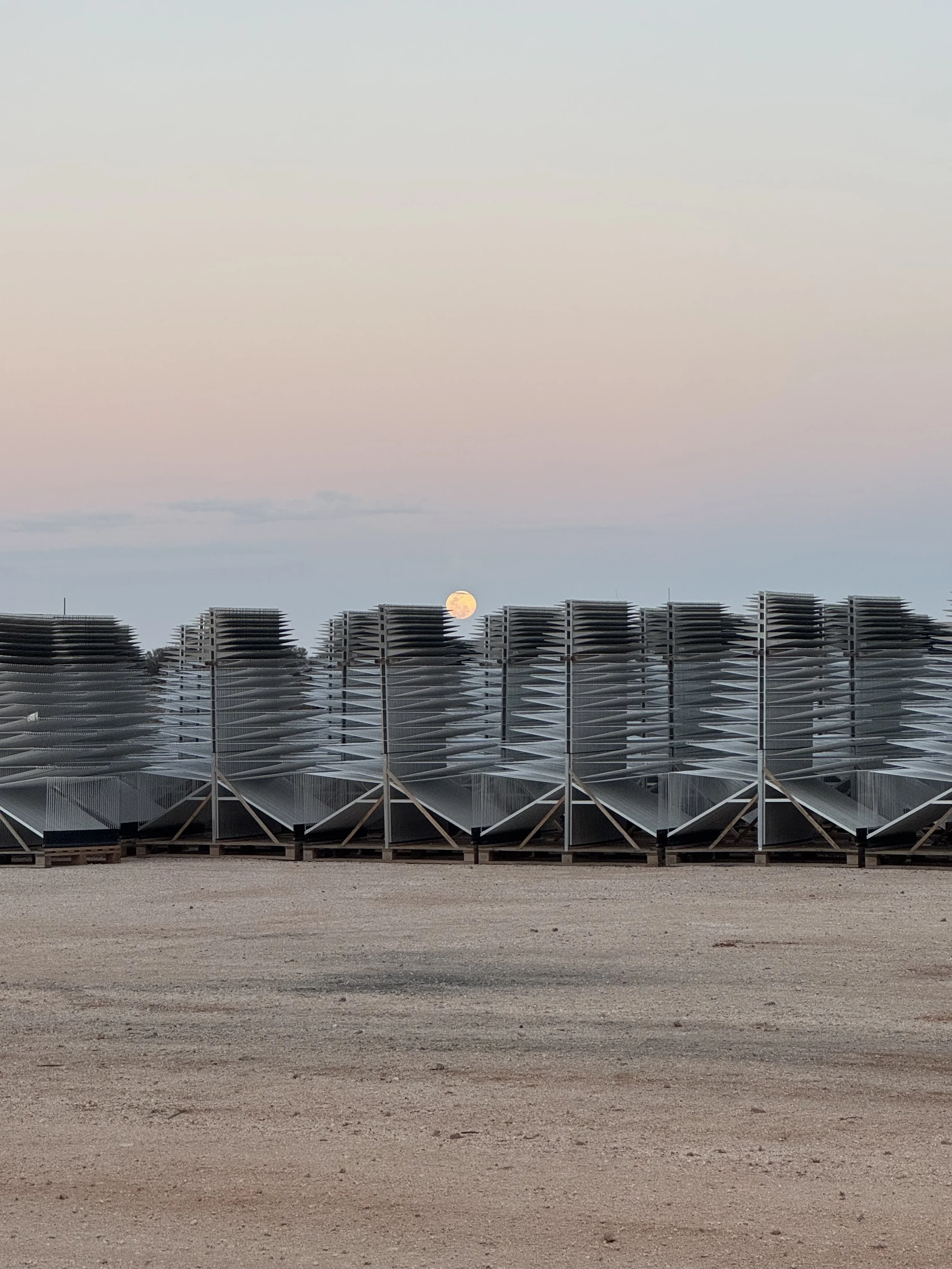 Sunset over a desert landscape with a full moon low on the horizon, and stacked metal structures in the foreground.