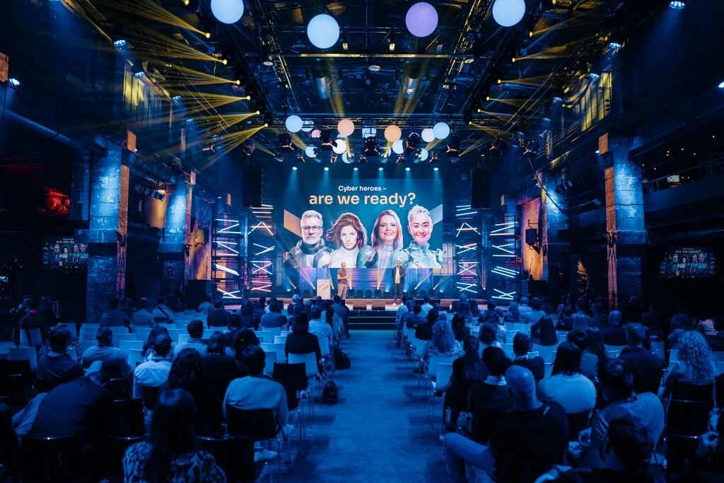 Large audience attending a conference with a stage featuring a digital screen displaying four cyber heroes and the question 'are we ready?'. The stage is decorated with neon lights and geometric designs, with lighting effects creating a vibrant atmos