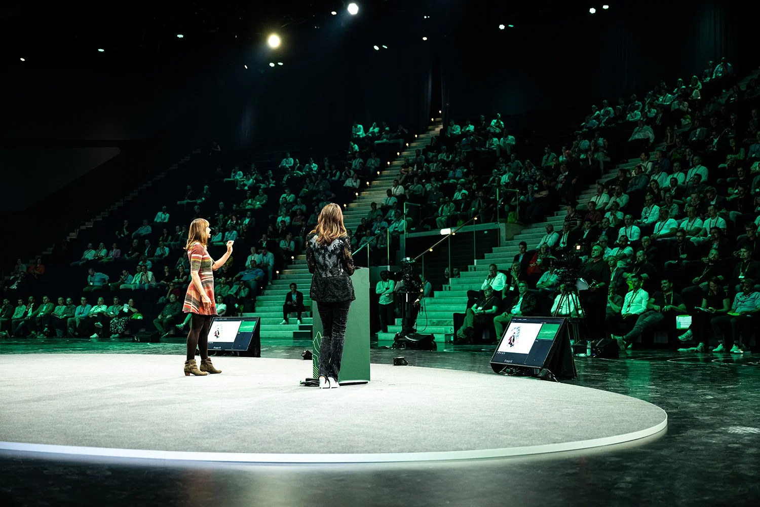 Two women presenting on stage to a large seated audience in a dark auditorium.