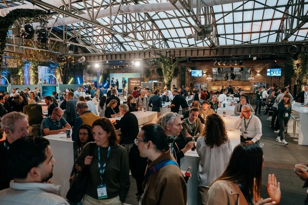Large indoor networking event or conference with many people interacting at booths and tables, decorated with plants and industrial-style architecture.