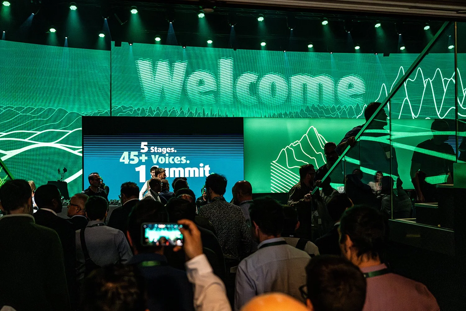 Conference scene with a large digital screen displaying the words 'welcome' and a smaller screen with text '5 stages, 45+ voices, 1 summit'. Attendees are gathered in front, some taking photos.