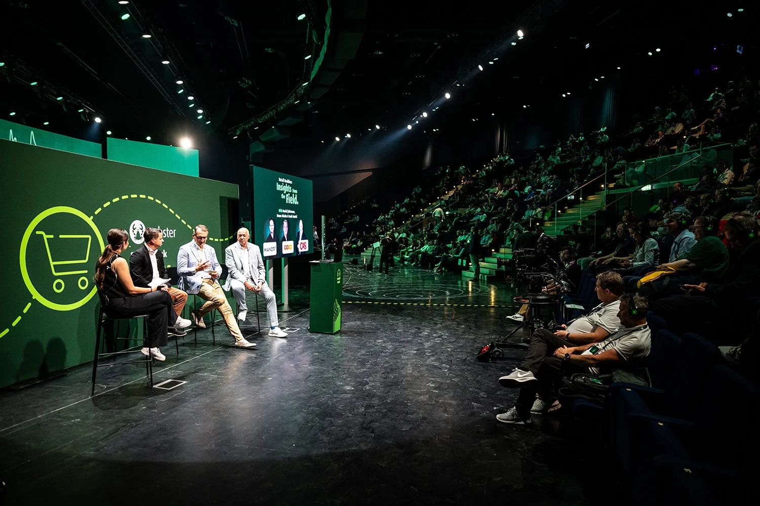 Panel discussion at a conference in a dark auditorium with an audience, featuring four speakers seated on stage with a large green screen behind them displaying logos and text.