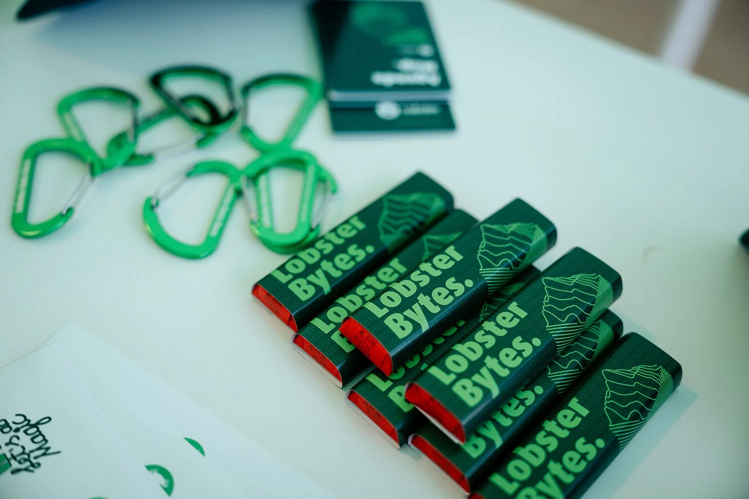 Multiple green and black "Lobster Bytes" energy bars lined up on a white surface with green carabiners and a dark green box in the background.