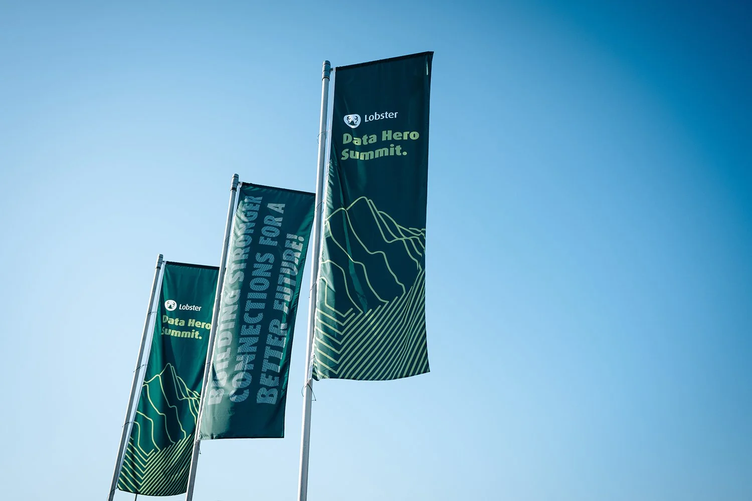 Three green flags with mountain graphics and text, positioned against a clear blue sky. The flags read "Lobster Data Hero Summit" and "Building Stronger Connections for a Better Future."
