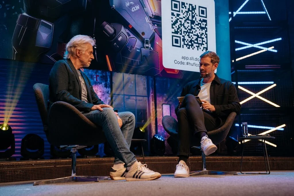 Two men sitting on stage in conversation with a digital background and a large screen displaying a QR code.