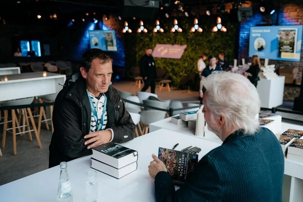 An author signing a book for a man at a book launch event, with books and a water bottle on the white table, in a modern, well-lit venue with other attendees and a presentation screen in the background.