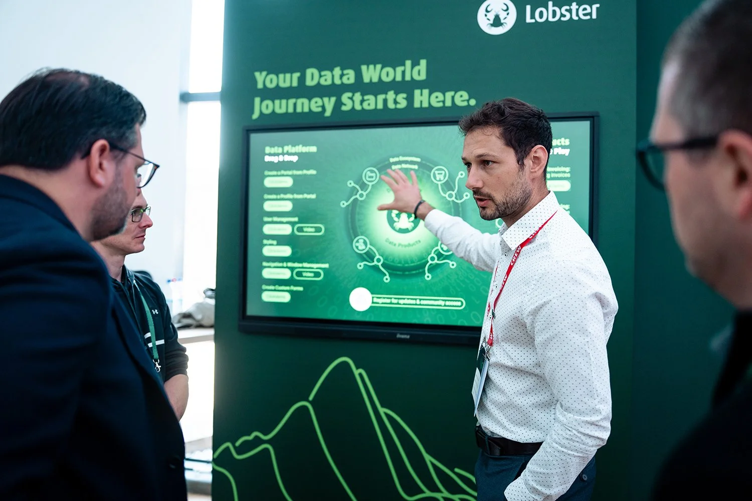 A man in a white shirt and explains data on a digital display to a group of people at a conference. The display reads 'Your Data World Journey Starts Here' and has a logo of a lobster.