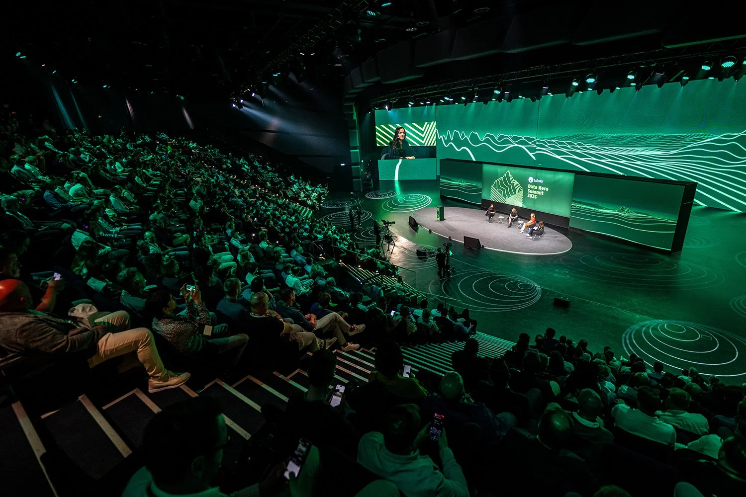 A large audience attending a conference or summit in an auditorium with a stage featuring a panel of four speakers. The stage has a digital display with green graphics and the text 'Data Hero Summit 2025'.