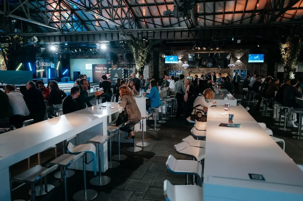 Indoor event space with high ceilings, featuring a stage at the front with screens and a sign that reads 'Ready to level up?', surrounded by seated attendees and modern white bar-style tables and chairs.