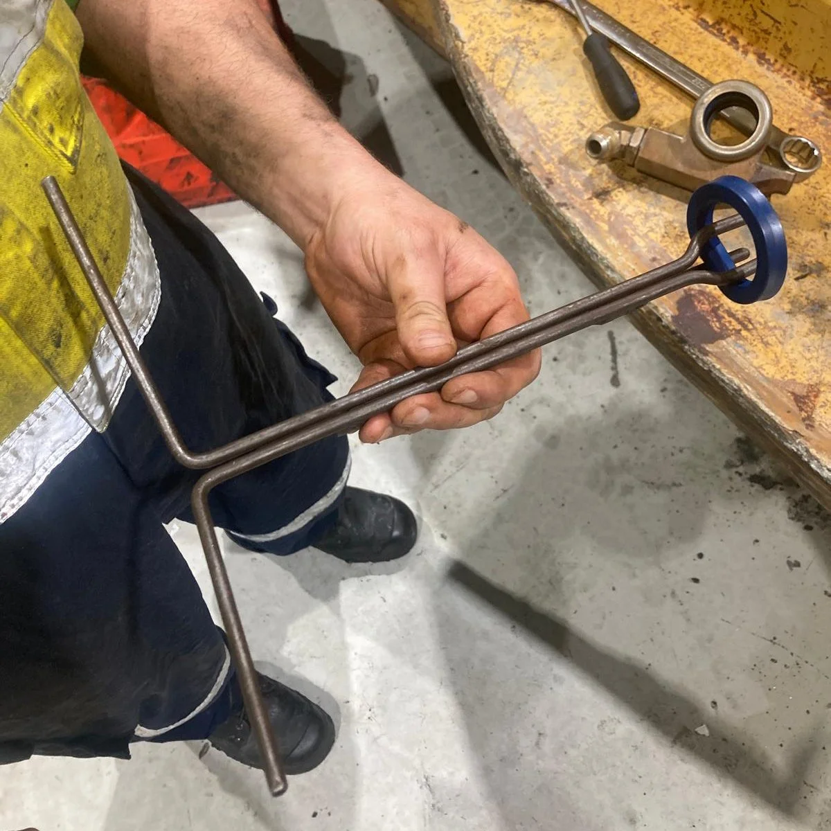 A worker holding a metal tool with a circular end, standing next to a workbench with various tools, in an industrial or workshop setting.