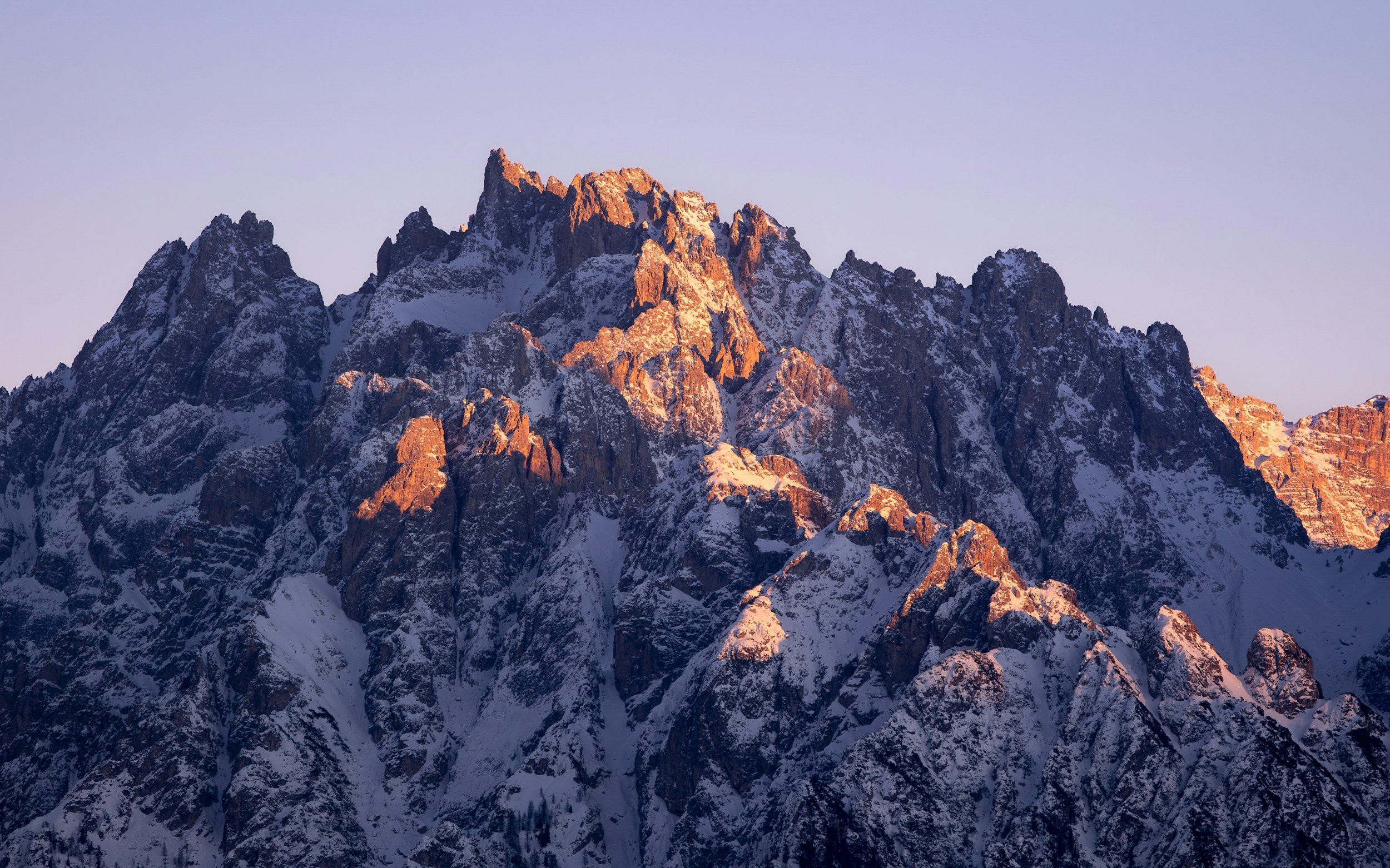Snow-capped rugged mountain peaks at sunset with pink-tinged rocks and a clear blue sky.