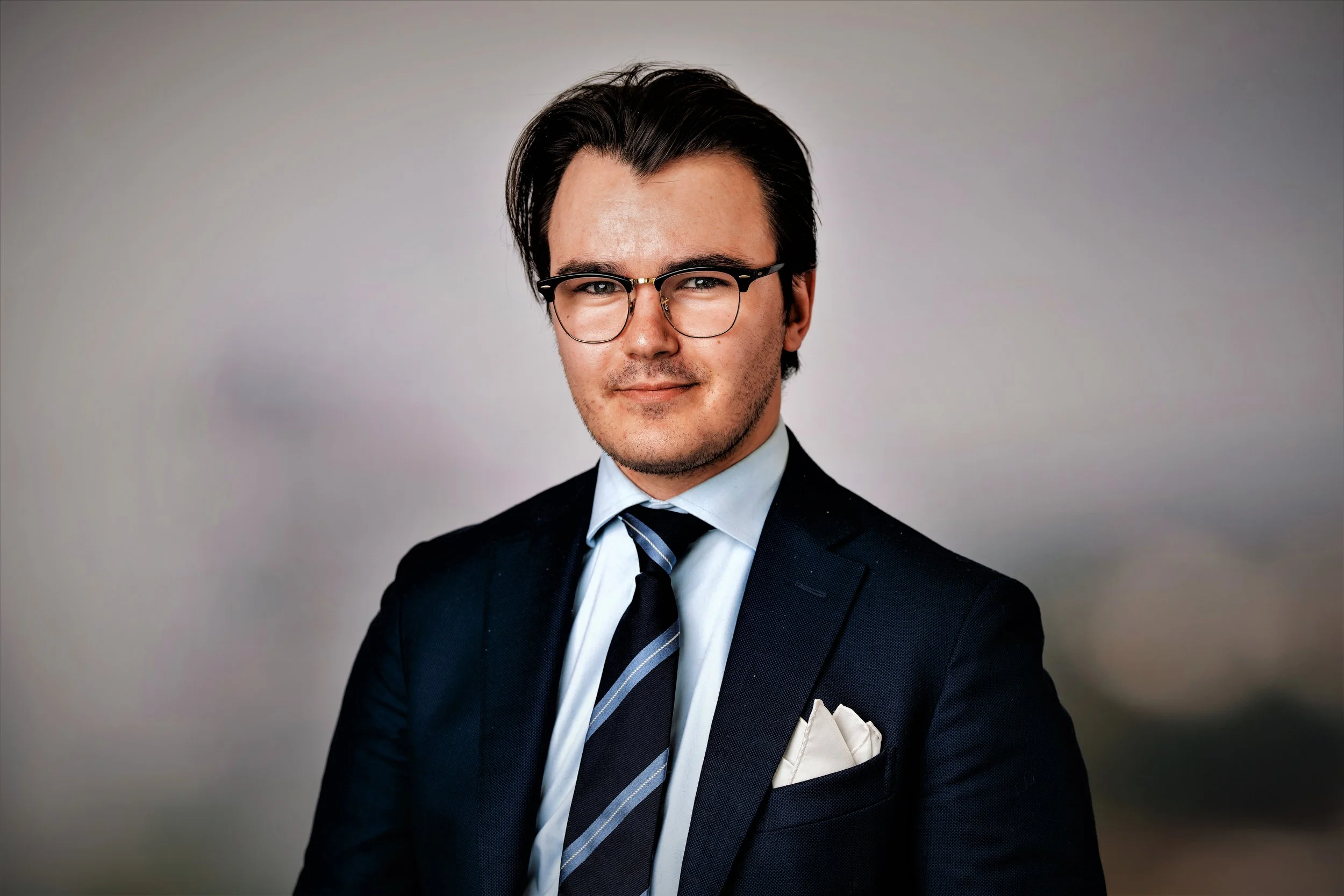 Portrait of a young man in a business suit with glasses, white shirt, striped tie, and pocket square against a neutral background.