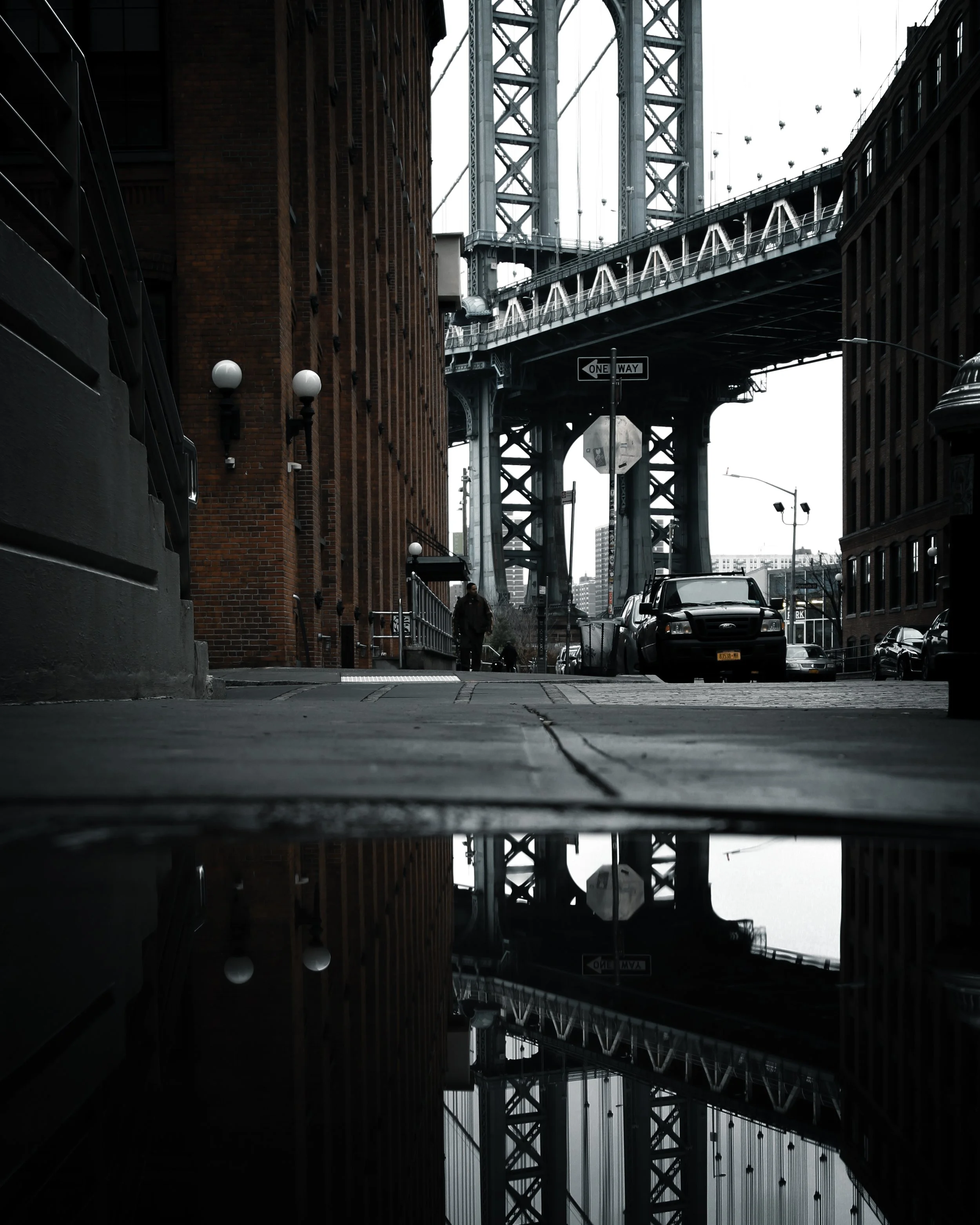 Urban street scene with a bridge overhead, cars parked along the street, a person walking, brick buildings, and a puddle reflecting the bridge and buildings.
