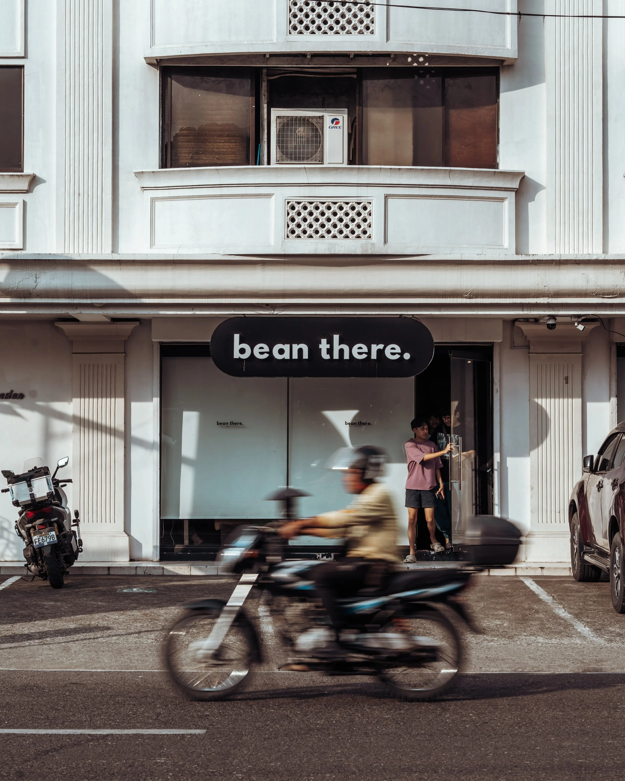 storefront with a black sign reading 'bean there.', a person on a motorcycle passing by, and people entering the store.