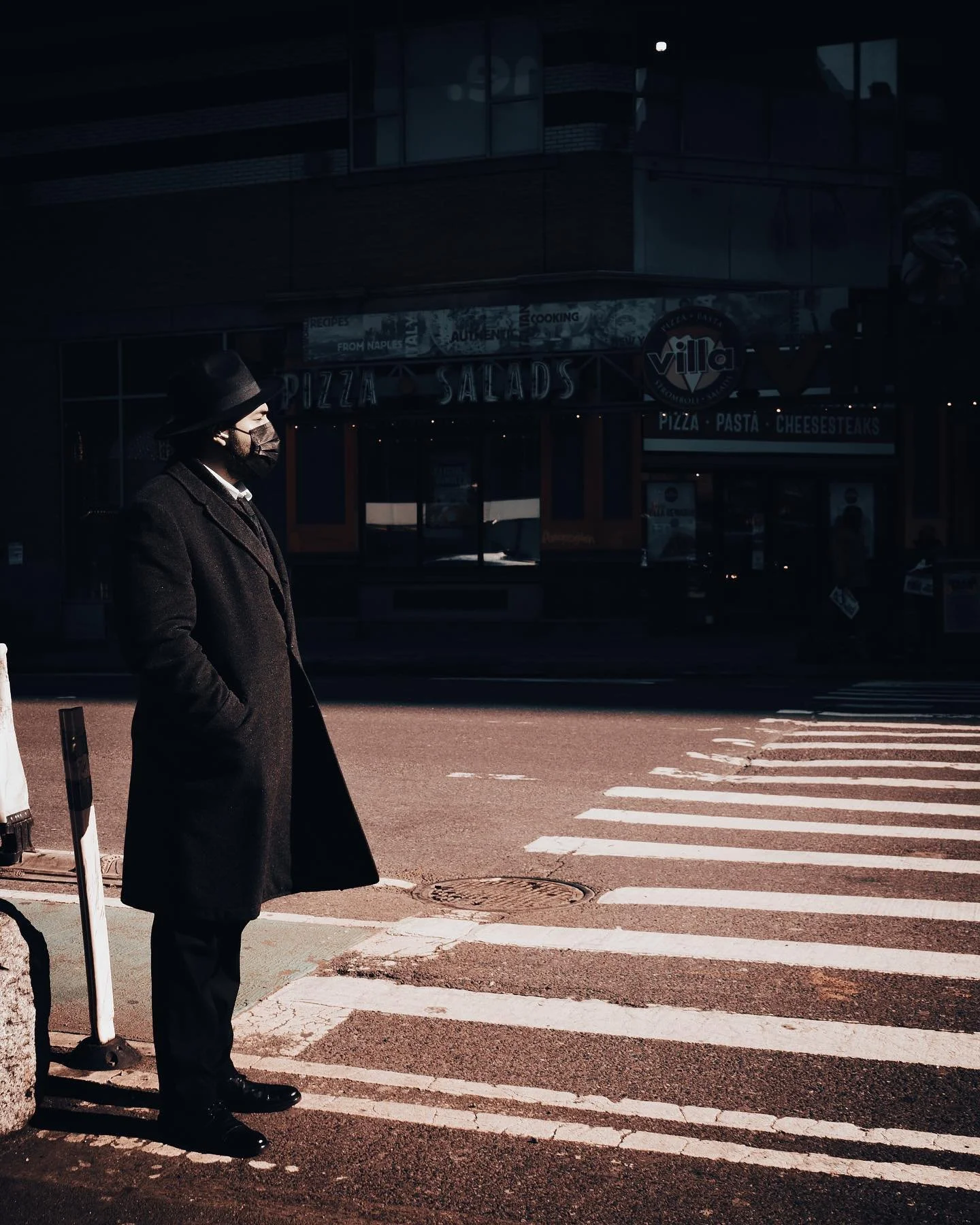 A person wearing a long coat, hat, and face mask standing on a crosswalk on a city street, with storefronts in the background.