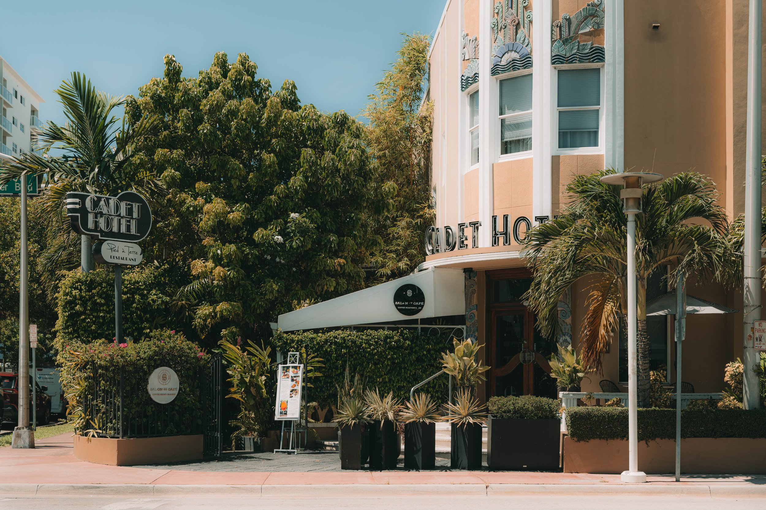 Exterior view of the Cadet Hotel with a sign, entrance, palm trees, and outdoor seating area in a sunny, tropical setting.