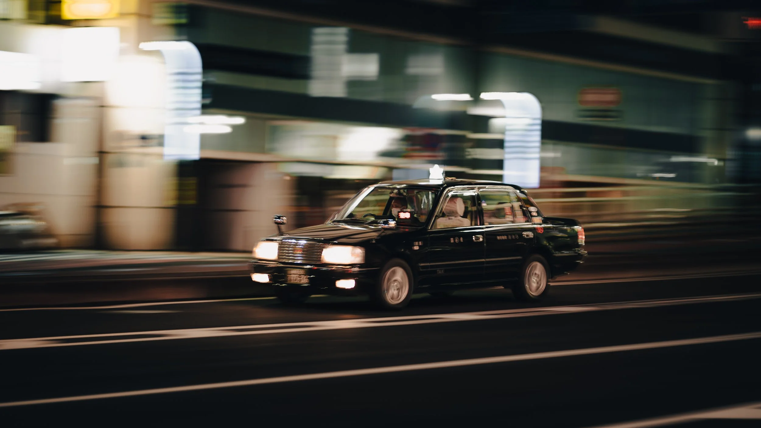 A black taxi cab driving on a city street at night with blurred lights and buildings in the background.