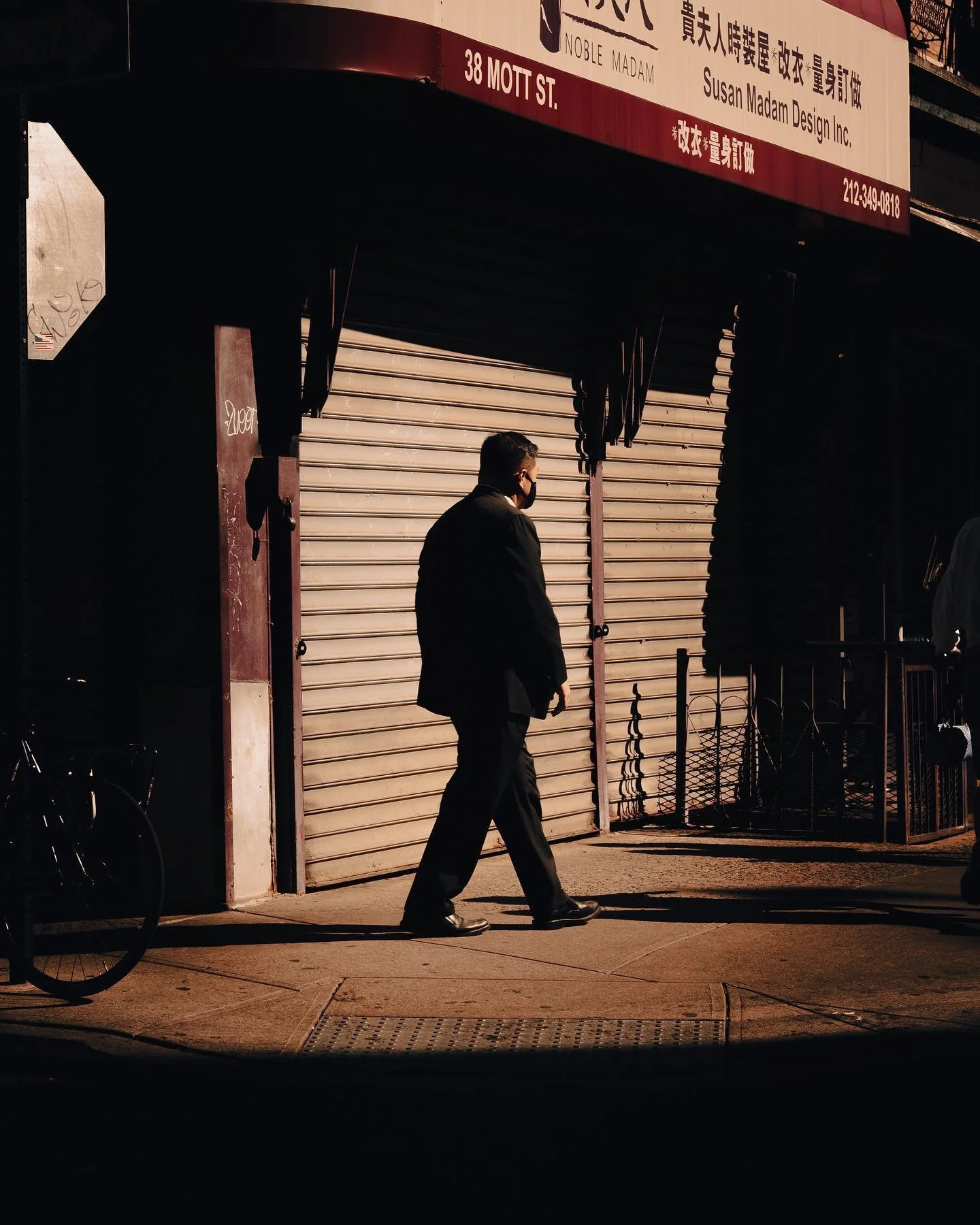 A man in a black suit and face mask walking on the sidewalk in front of a closed storefront with a metal shutter, illuminated by streetlights at night.