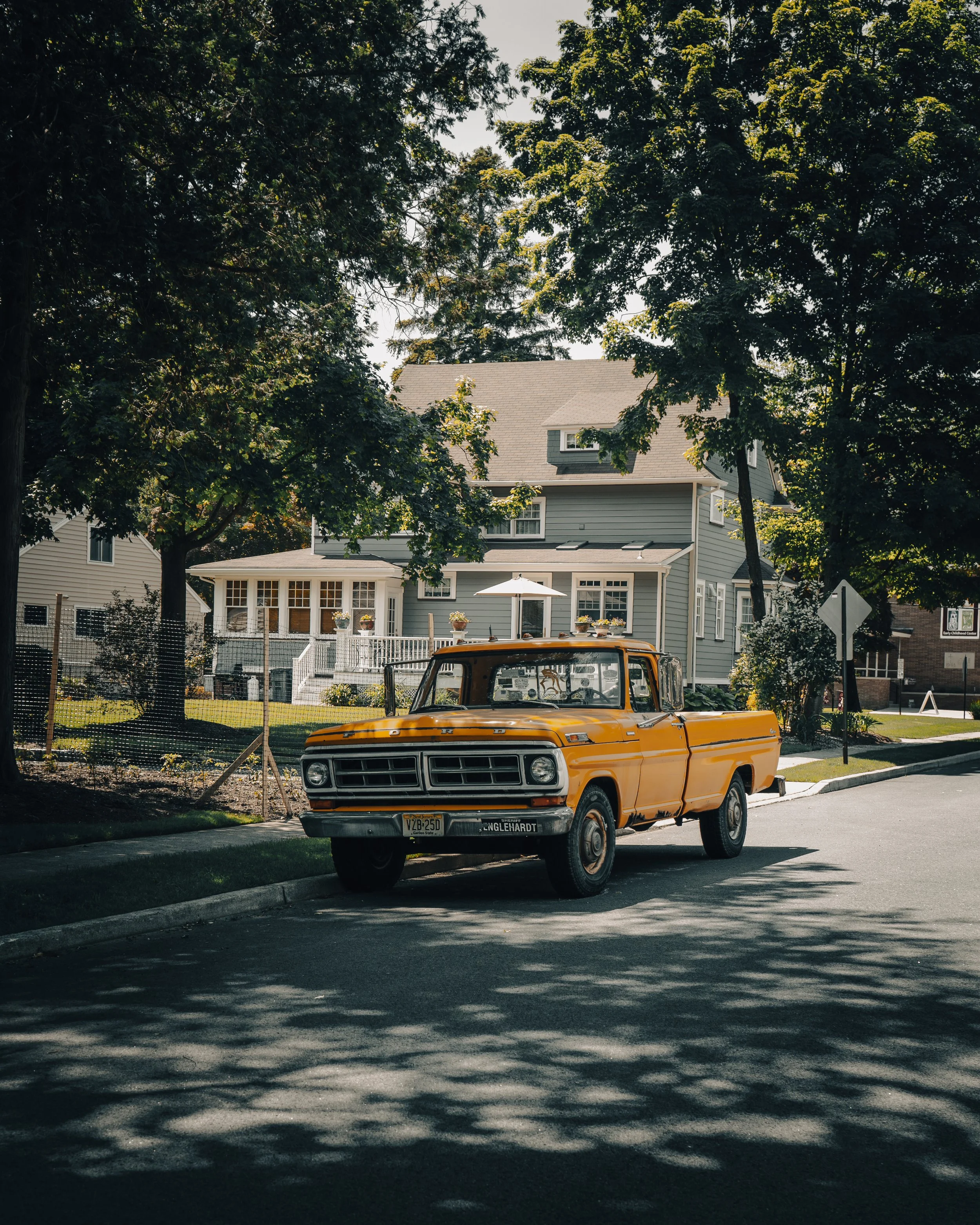 Orange vintage pickup truck parked on a residential street with trees and houses in the background.