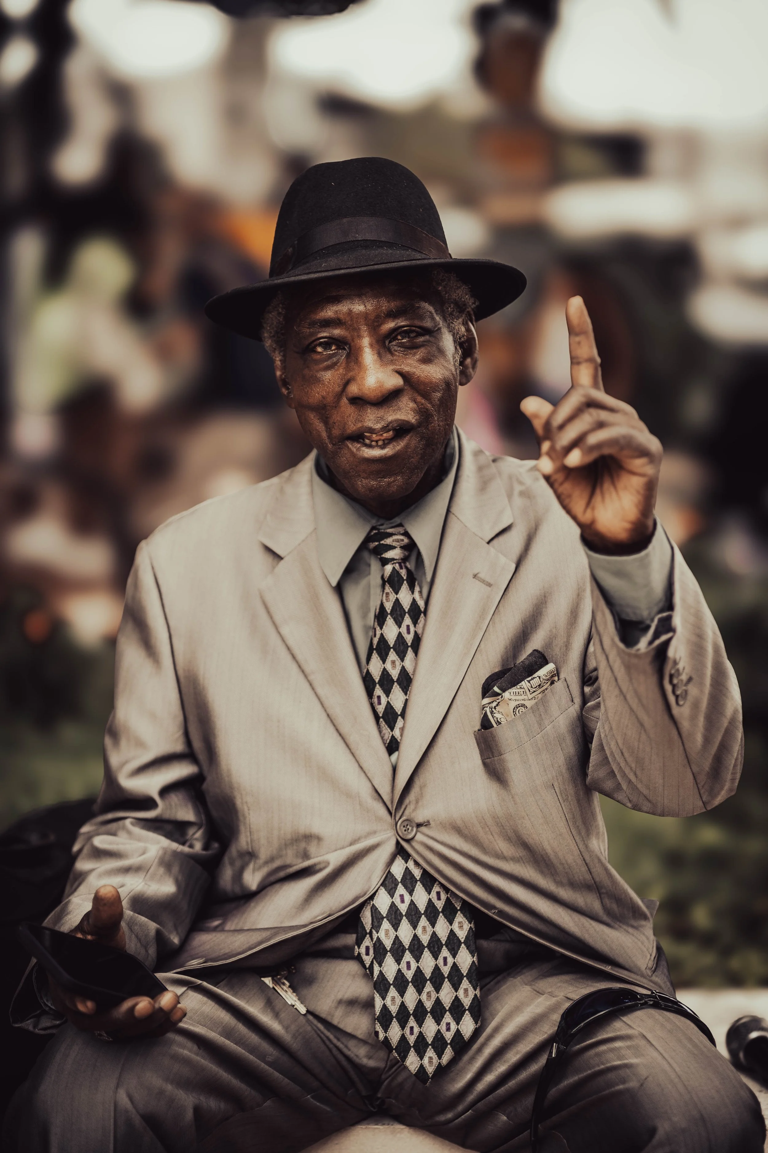 An elderly man in a beige suit and fedora hat sitting outdoors, holding a smartphone in his left hand and gesturing with his right index finger raised, with a blurred crowd in the background.