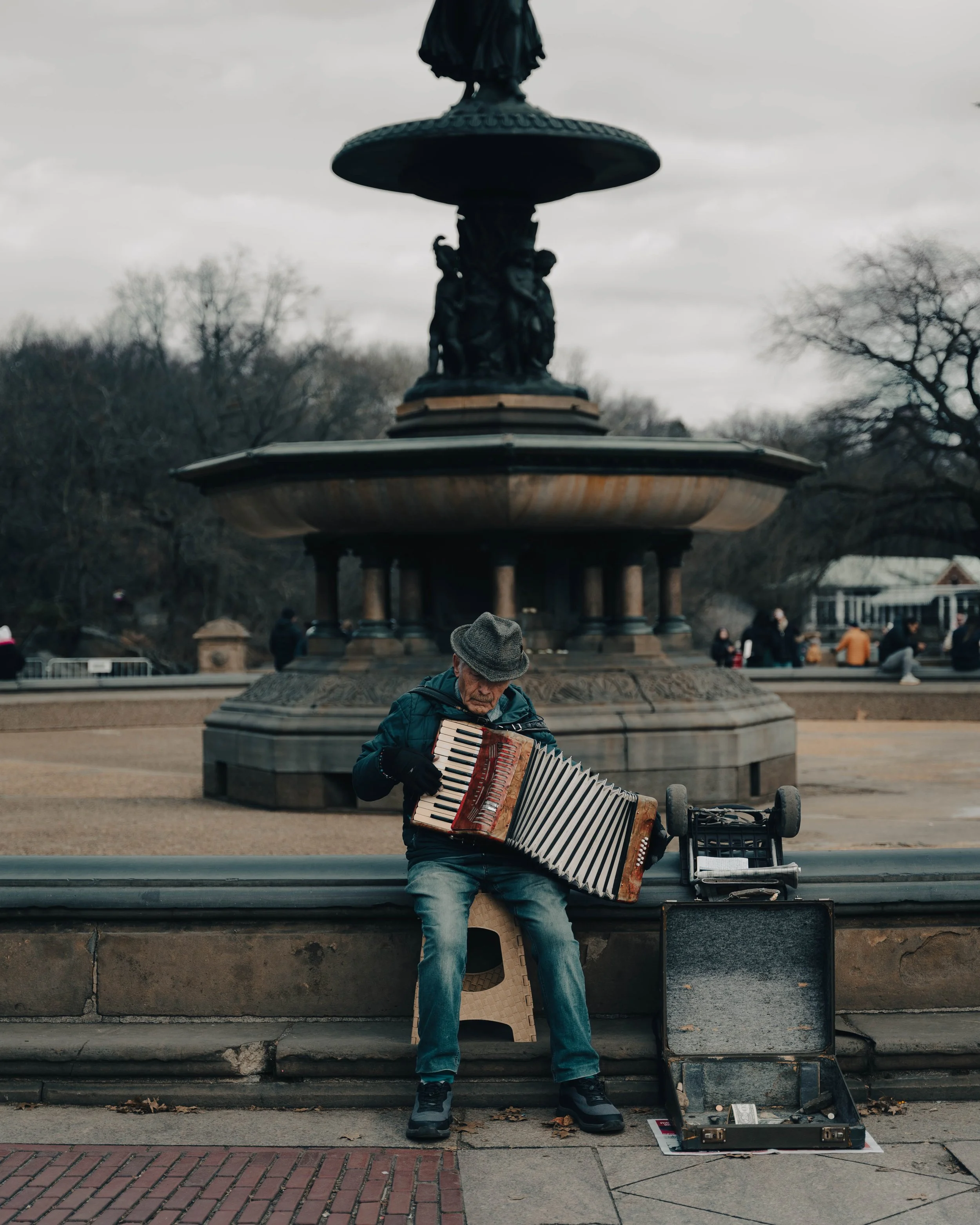 An elderly man playing an accordion on a city sidewalk in front of a fountain, with other people in the background on a cloudy day.