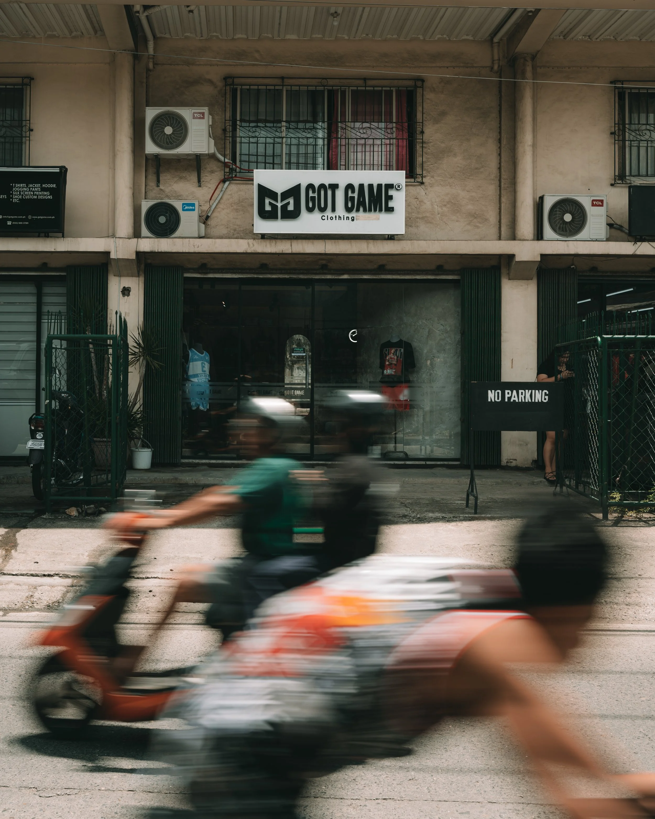 Blurred person riding a motorcycle passing in front of a storefront with a sign that reads 'GOT GAME Clothing' and a 'NO PARKING' sign near a gate.