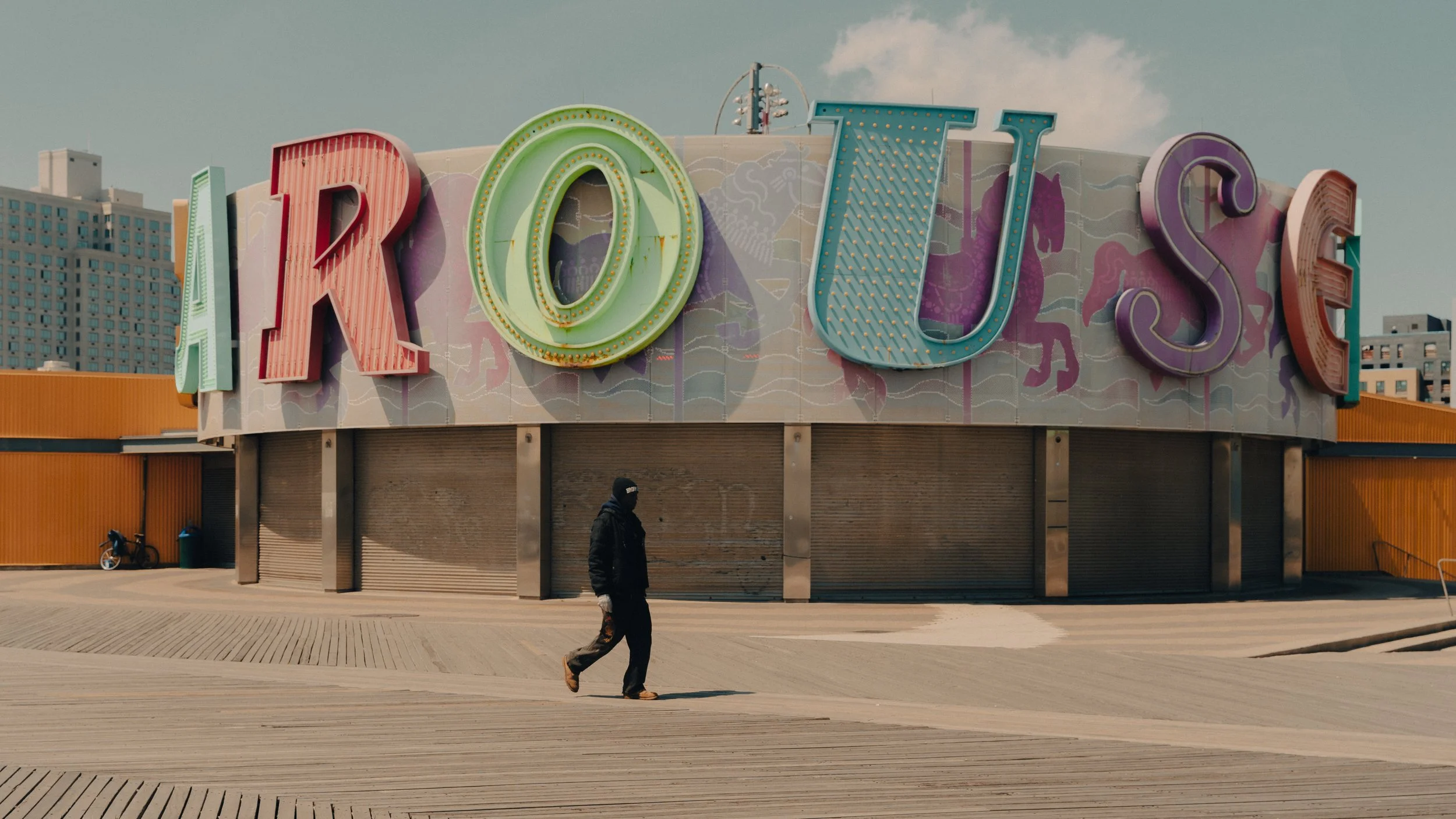 A person walking past an amusement park entrance with large colorful letters spelling 'AROUS'. The background shows a city skyline with tall buildings.