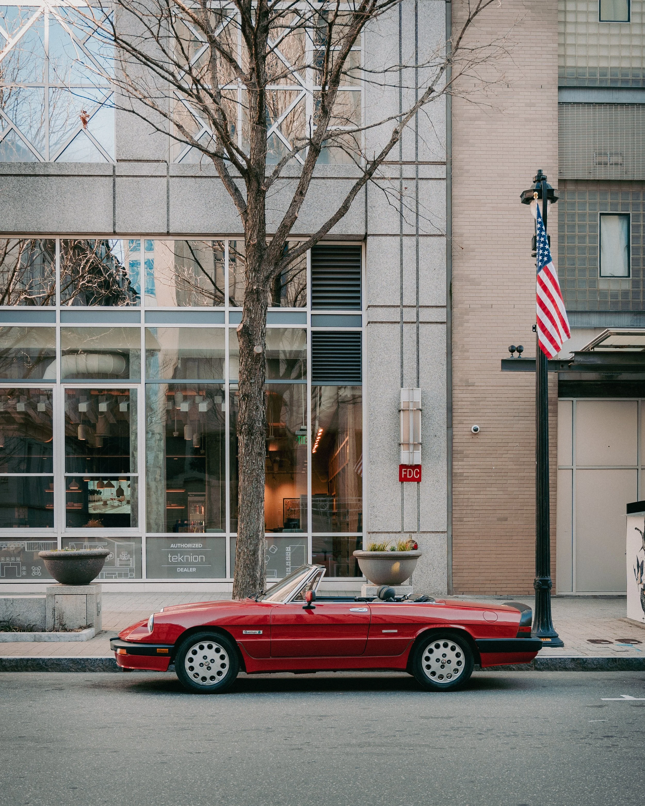 Red vintage convertible car parked on city street in front of a modern building with large glass windows. Tree with no leaves and an American flag on a pole also visible.