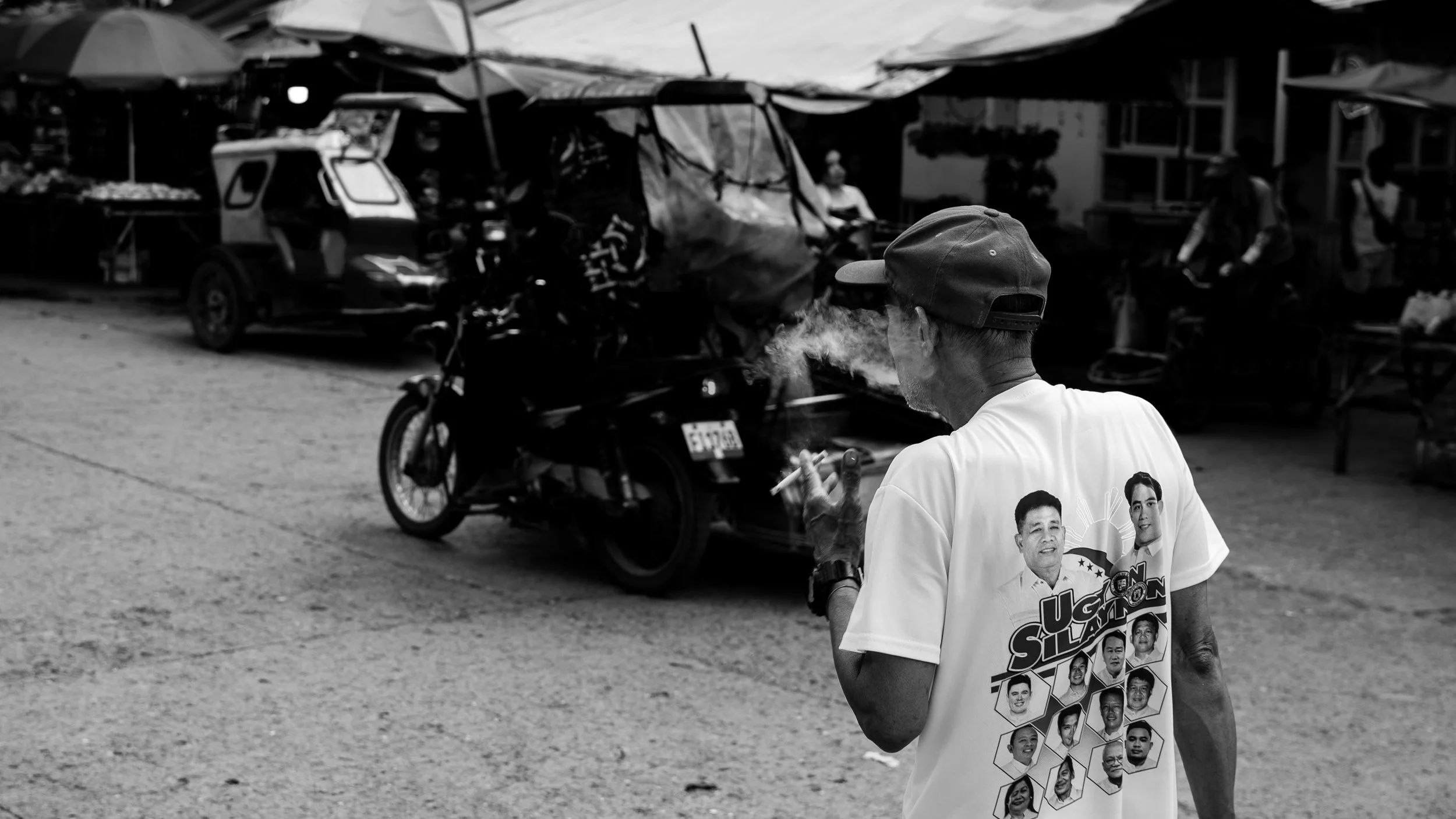 A man wearing a white T-shirt with political images and text, smoking and holding a cigarette in a busy outdoor market with vehicles and market stalls in the background.