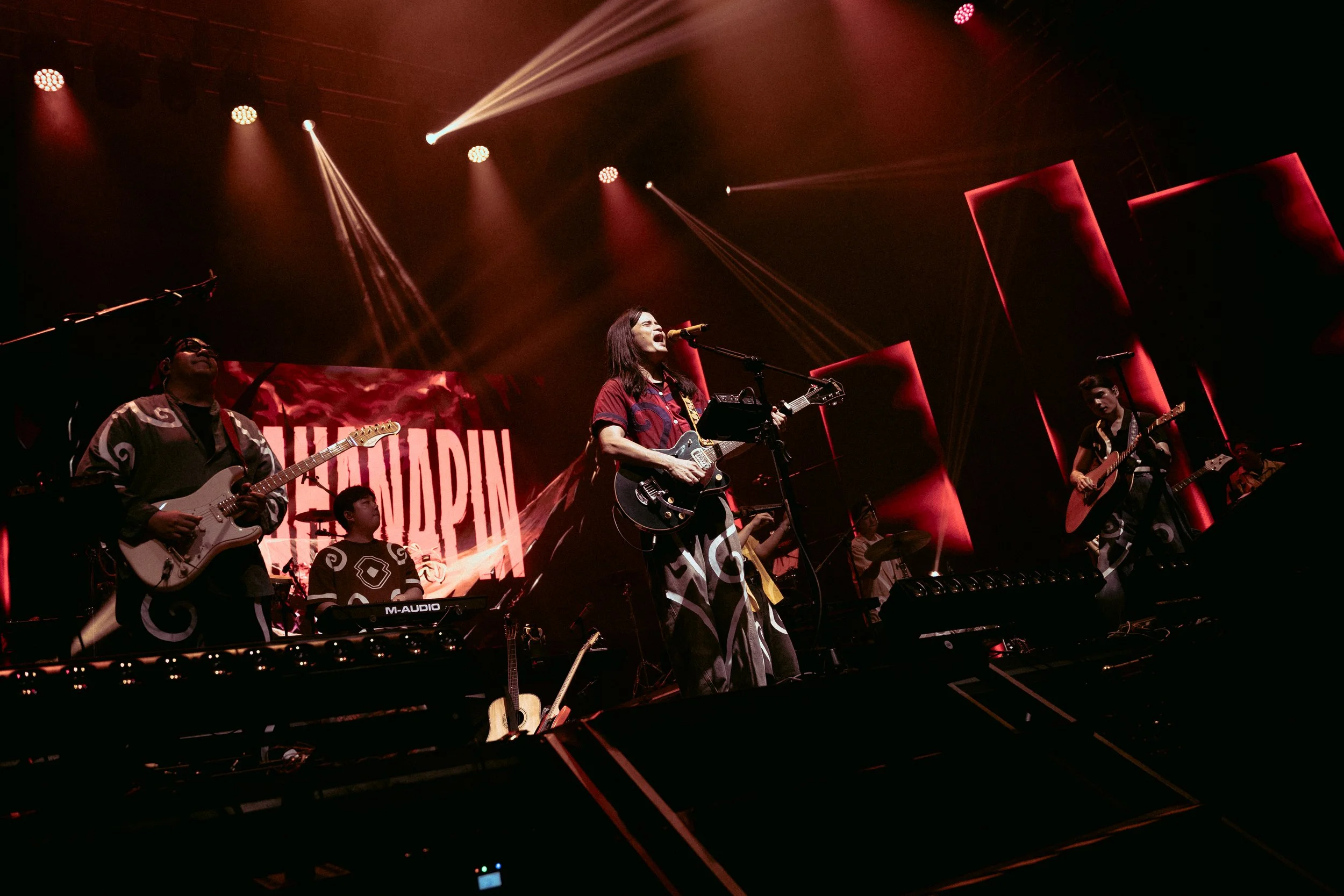 A musical band performing on stage with multiple musicians playing guitars and drums, illuminated by red stage lights, with a large screen displaying the word 'LADIN' in the background.