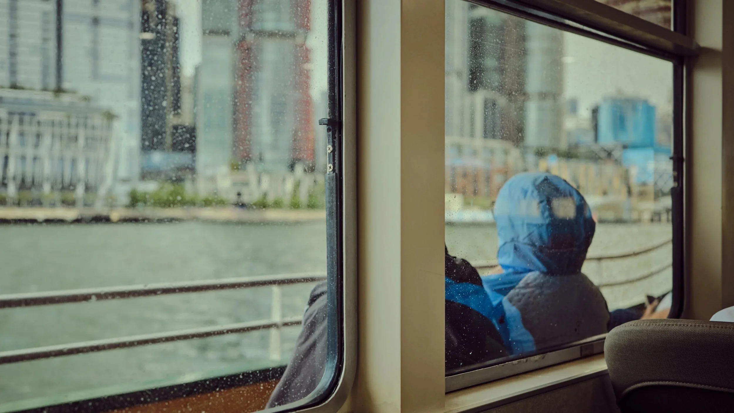 View from inside a bus showing a person in a blue hoodie sitting on the right side, looking out the window at a rainy cityscape with buildings and water.