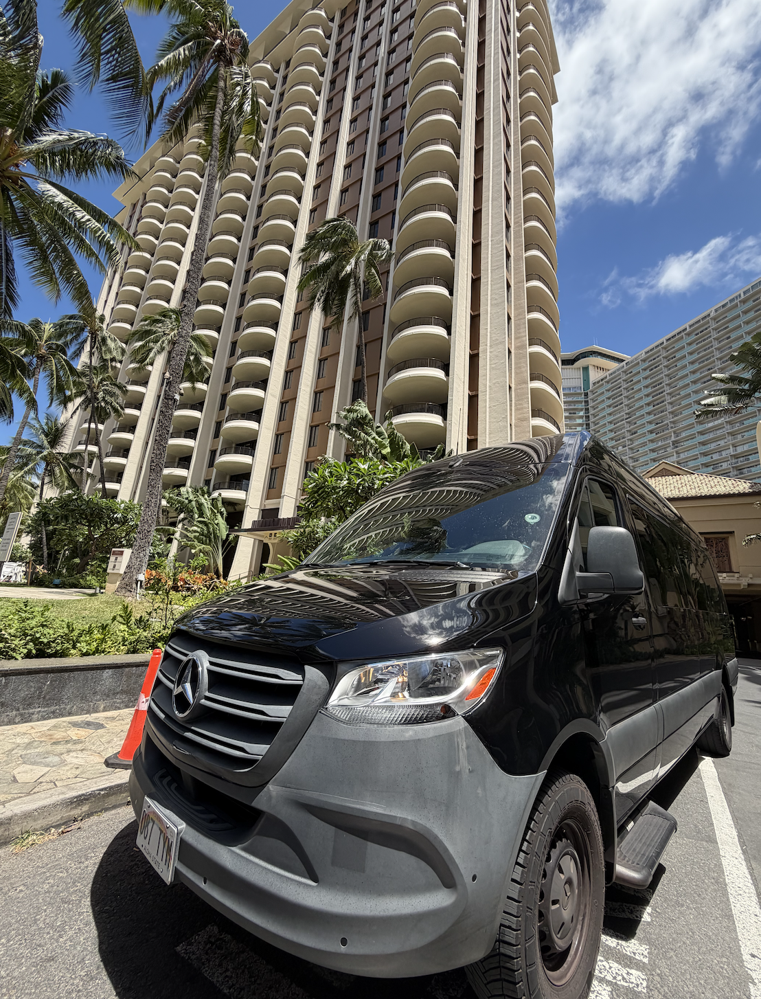 A black Mercedes-Benz van parked in front of a tall residential building with curved balconies, surrounded by palm trees, under a partly cloudy blue sky.