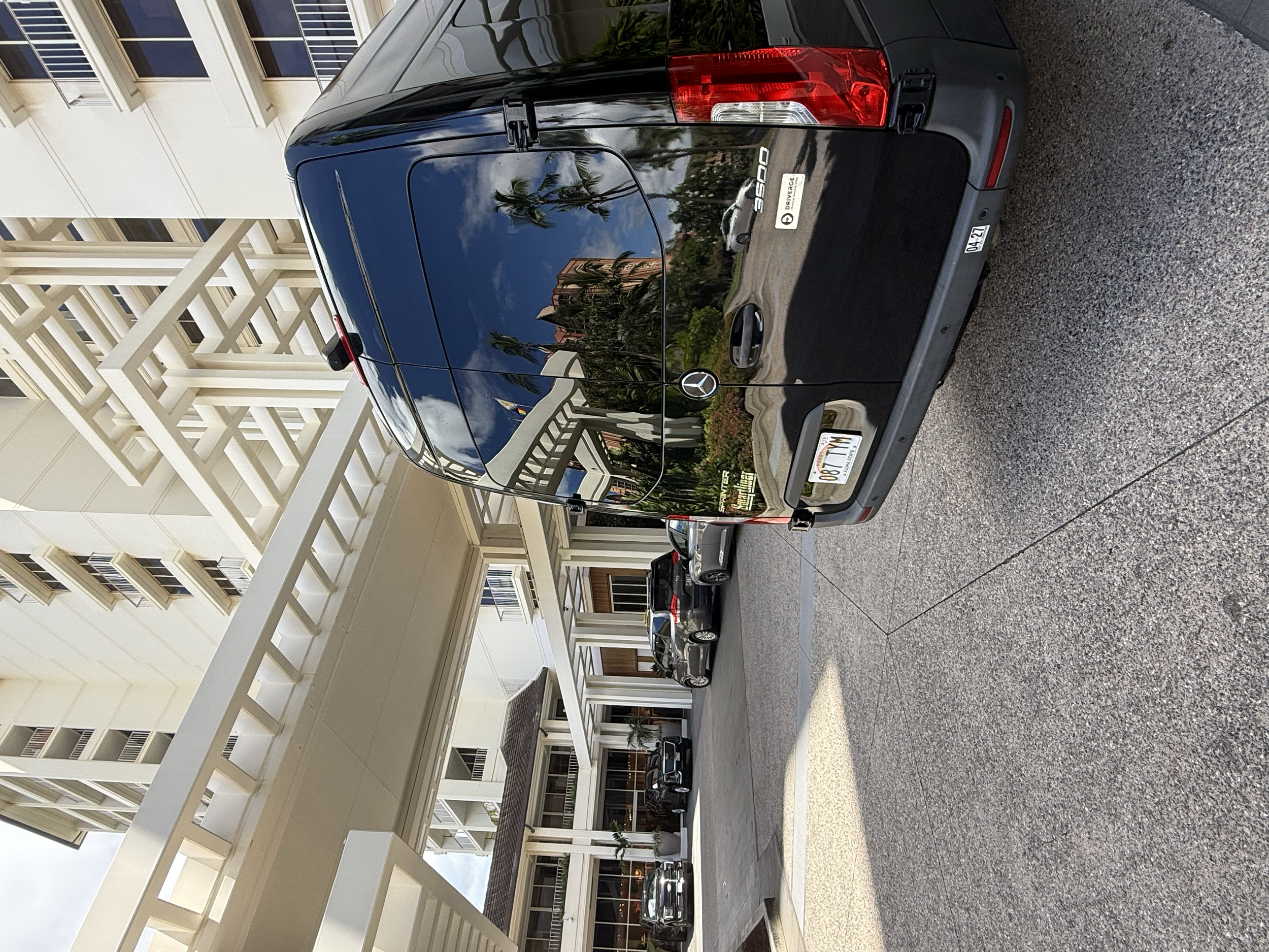 The image shows a black Mercedes-Benz G-Class SUV parked in front of a modern multi-story apartment building with white exterior walls and large windows. The car's surface reflects palm trees and the sky.