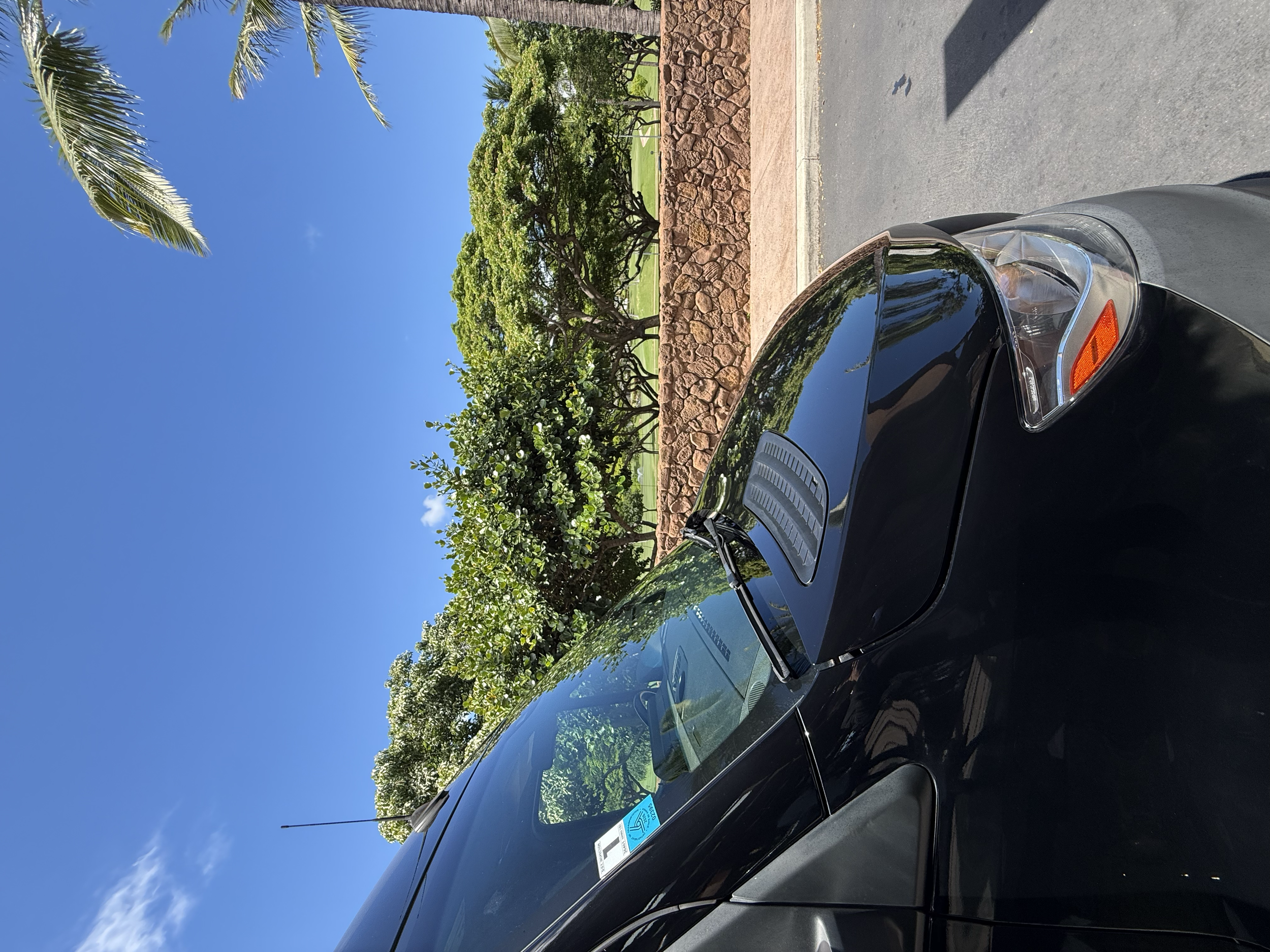 Close-up of a black car parked on a street with a stone wall, trees, and a blue sky in the background.