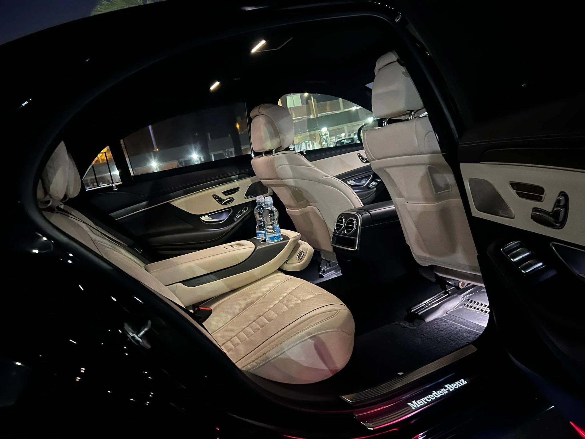 Interior of a black luxury car with beige leather seats, two water bottles on the center console, and ambient lighting, branded with Mercedes Benz.
