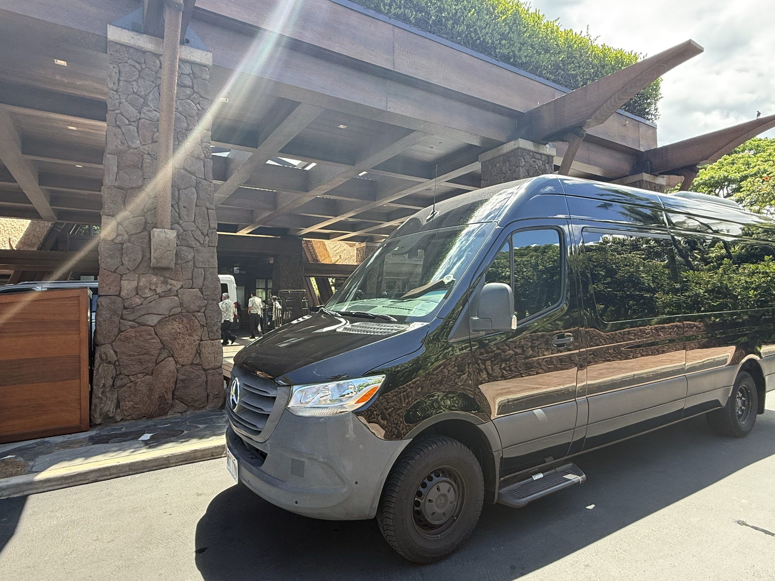 Black Mercedes-Benz van parked on the street in front of a building with stone columns and a wooden overhang, with trees and a partly cloudy sky in the background.