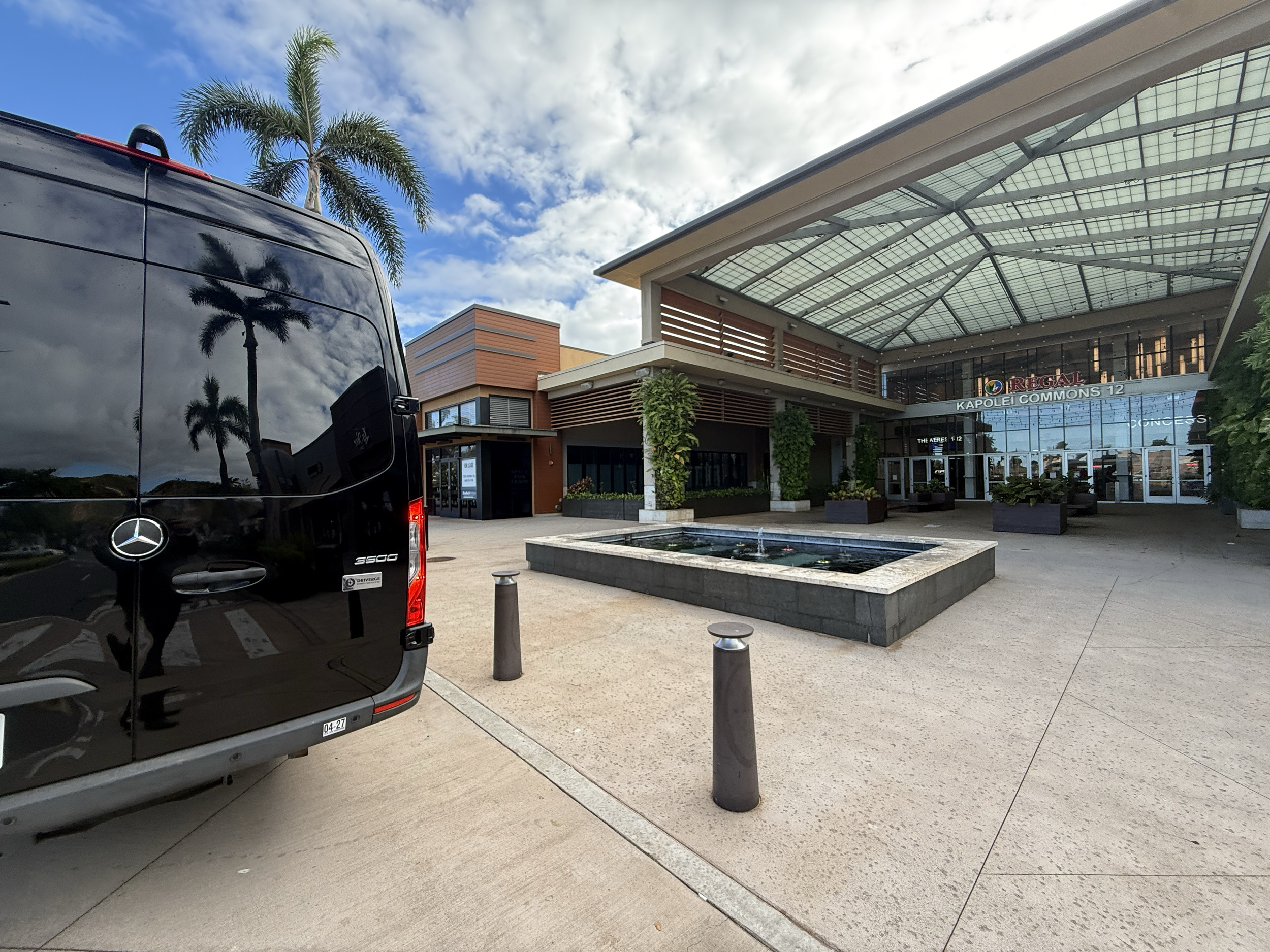 Exterior view of a modern shopping mall called 'Reagal' with palm trees, a water fountain, a glass canopy, and a parked Mercedes-Benz van.