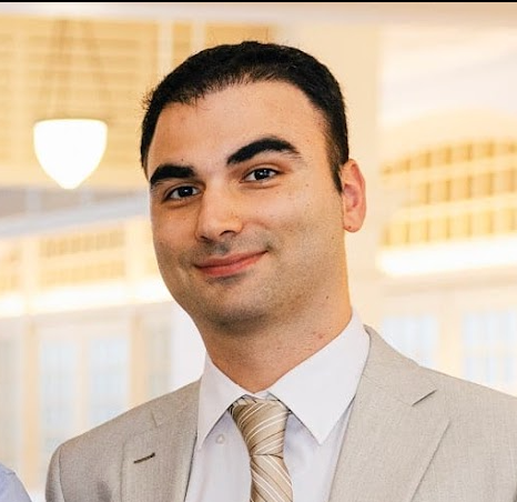 A man in a beige suit and tie smiling in an indoor setting with soft lighting and modern building windows in the background.
