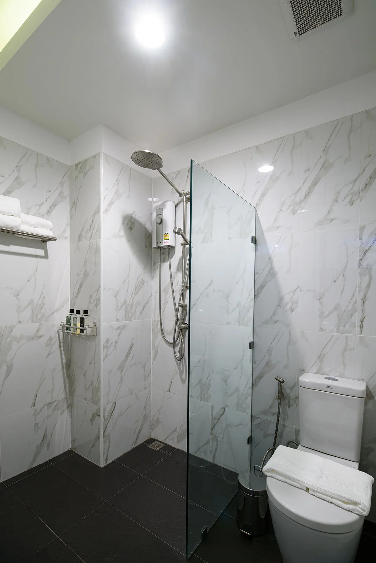 Modern bathroom with white marble walls, black floor tiles, a glass shower partition, a showerhead, a towel shelf, and a white toilet with a rolled towel on top.