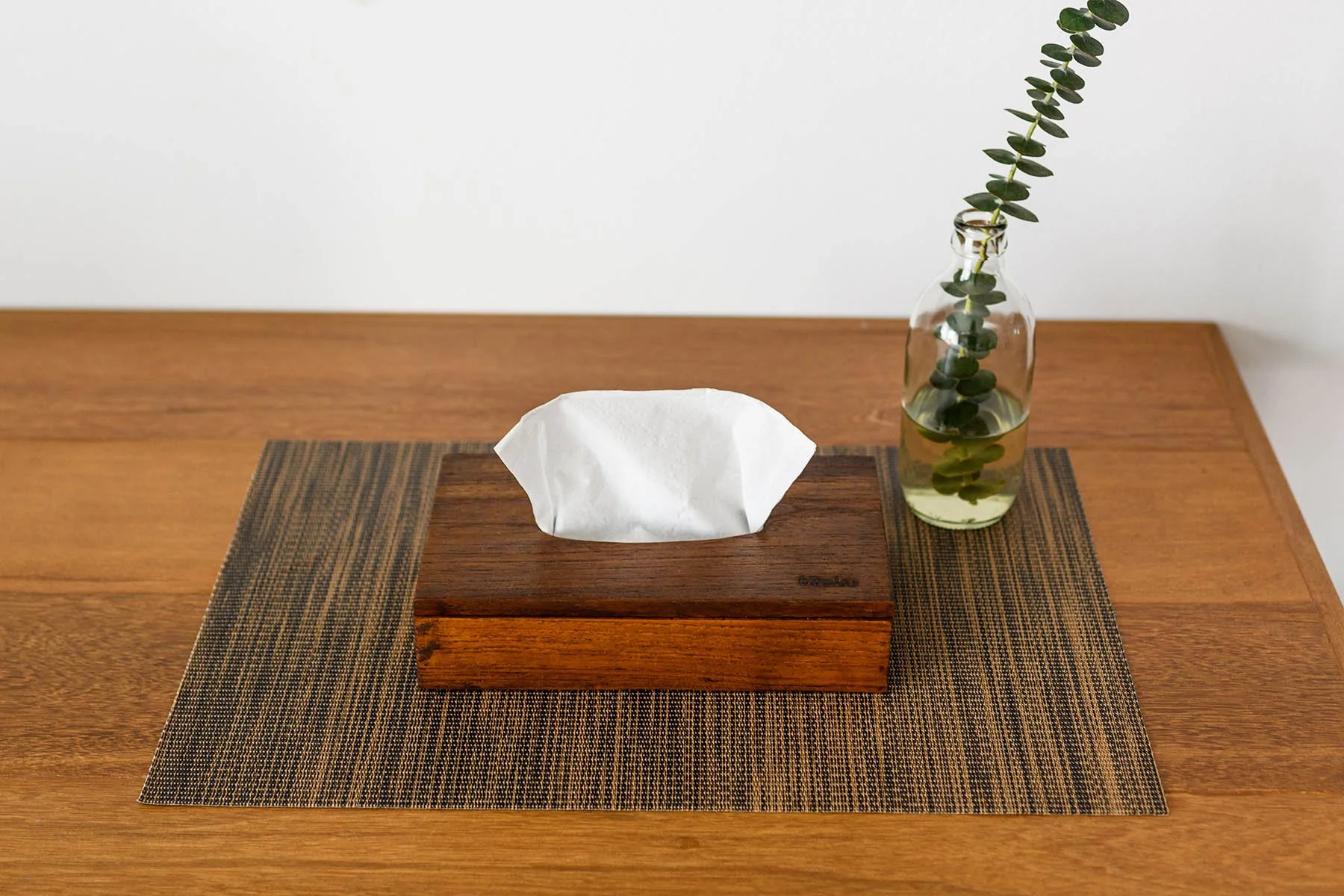 A wooden tissue box with a tissue protruding, placed on a woven placemat on a wooden table, with a glass bottle containing a branch of greenery to the right.