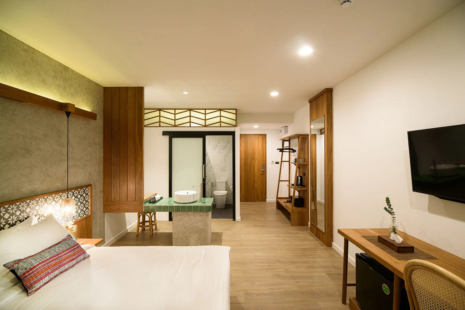 A hotel room with a bed, a floating nightstand with a lamp, a workspace, and a bathroom with a frosted glass sliding door, wood flooring, and modern decor.