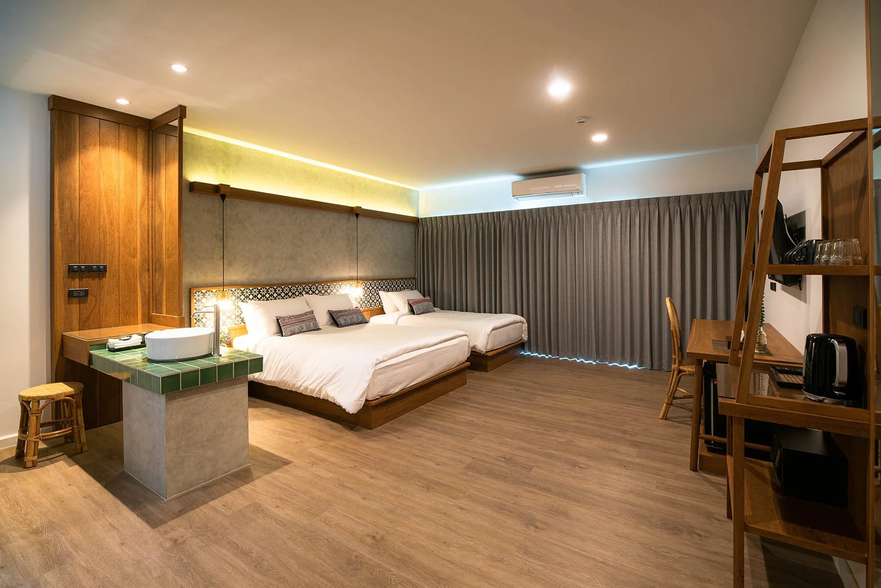 A modern hotel room with two beds, a wooden desk with a chair, a shelving unit with a TV, and a small green-tiled table with a sink. The room has wooden floors, grey curtains, and warm lighting.