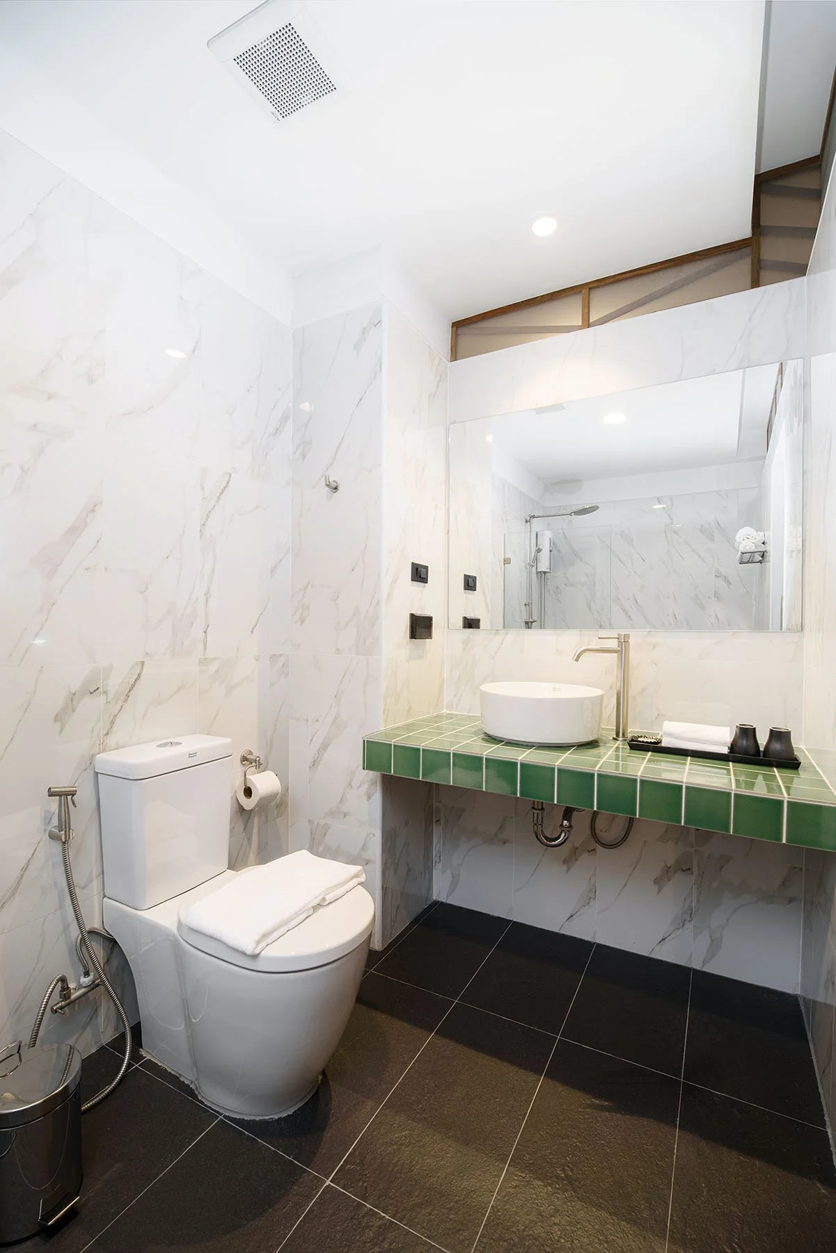 Modern bathroom with white marble walls, black floor tiles, a toilet with a towel on top, a green tiled countertop with a vessel sink, a large mirror, and a shower area with a glass door.