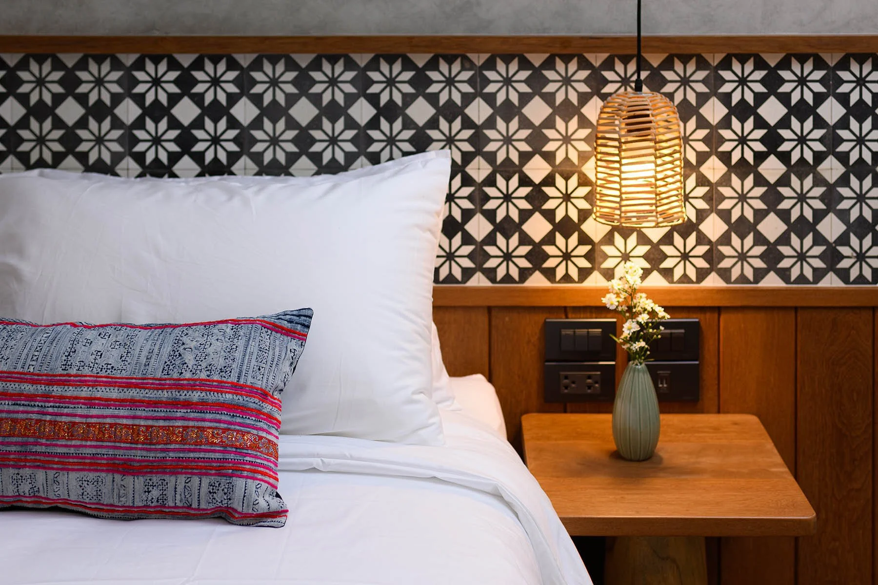 Close-up of a neatly made bed with white pillows and a decorative pillow, beside a wooden bedside table with a vase of white flowers and a hanging woven lampshade, against a wall with black and white geometric patterned tiles.