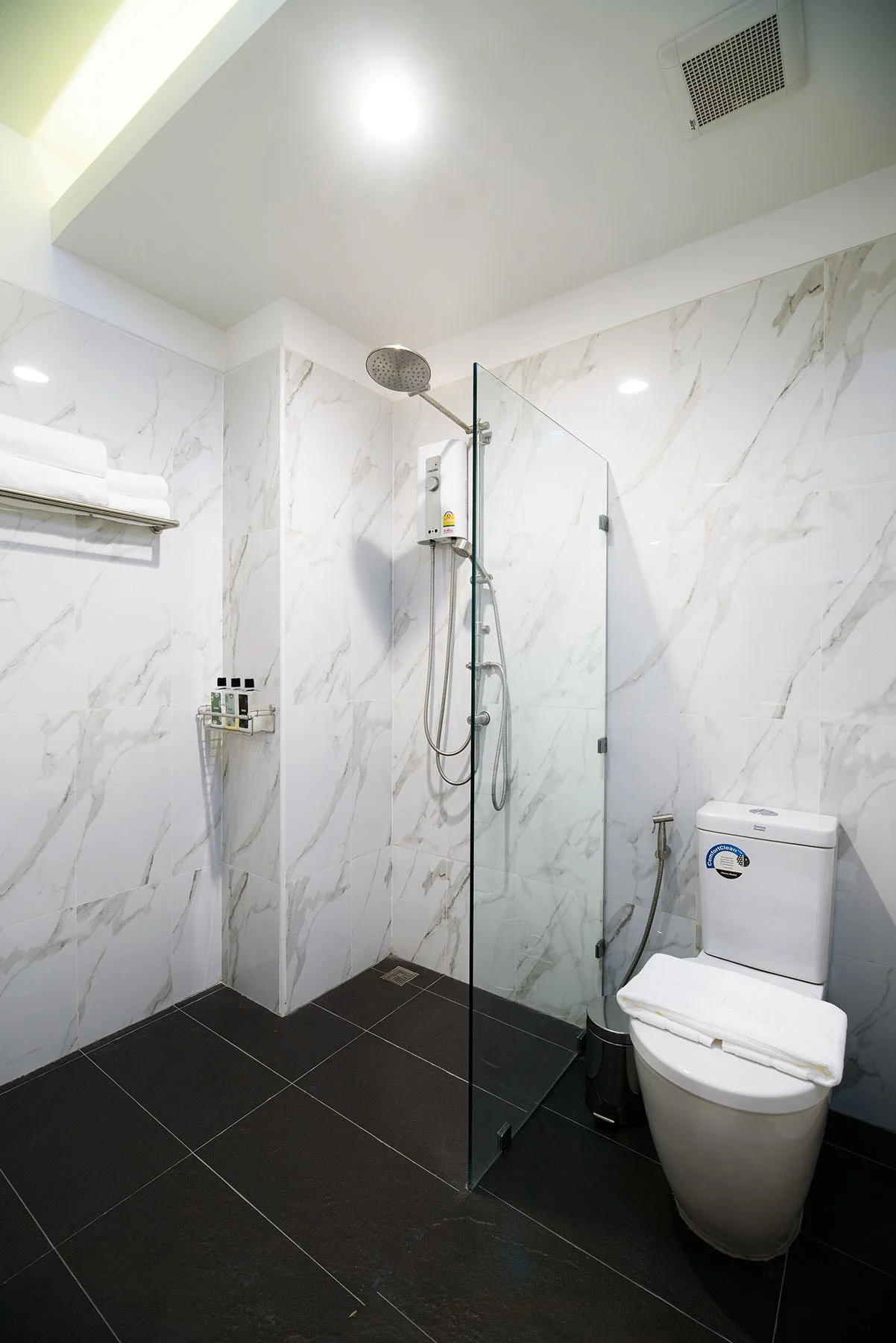 Modern bathroom with white marble walls, black tiled floor, glass shower partition, rainfall showerhead, toilet, and a shelf with towels