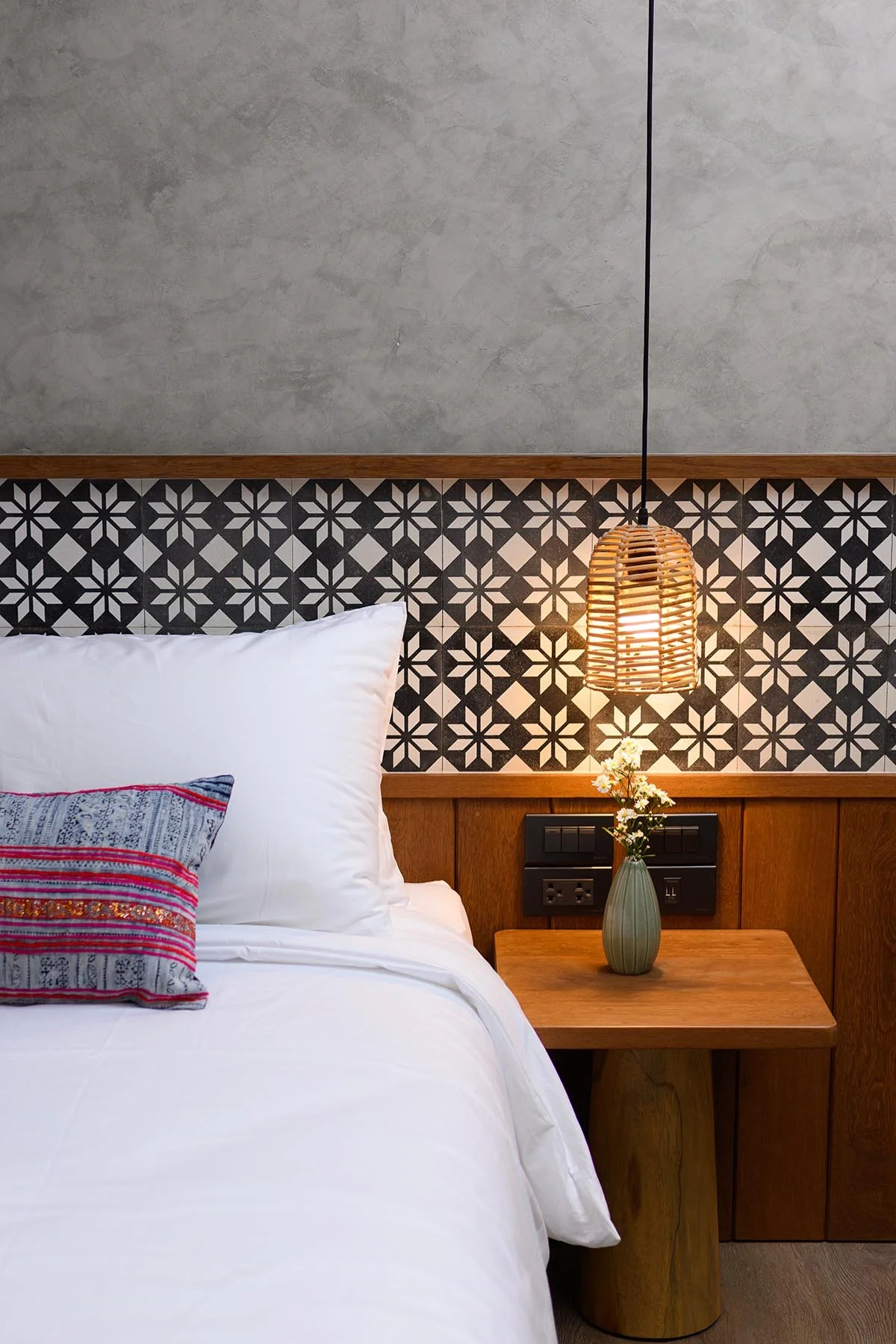 A bedroom corner with a white bed, colorful pillow, wooden bedside table, vase with flowers, textured gray wall, patterned black and white tile, and a hanging woven lamp.