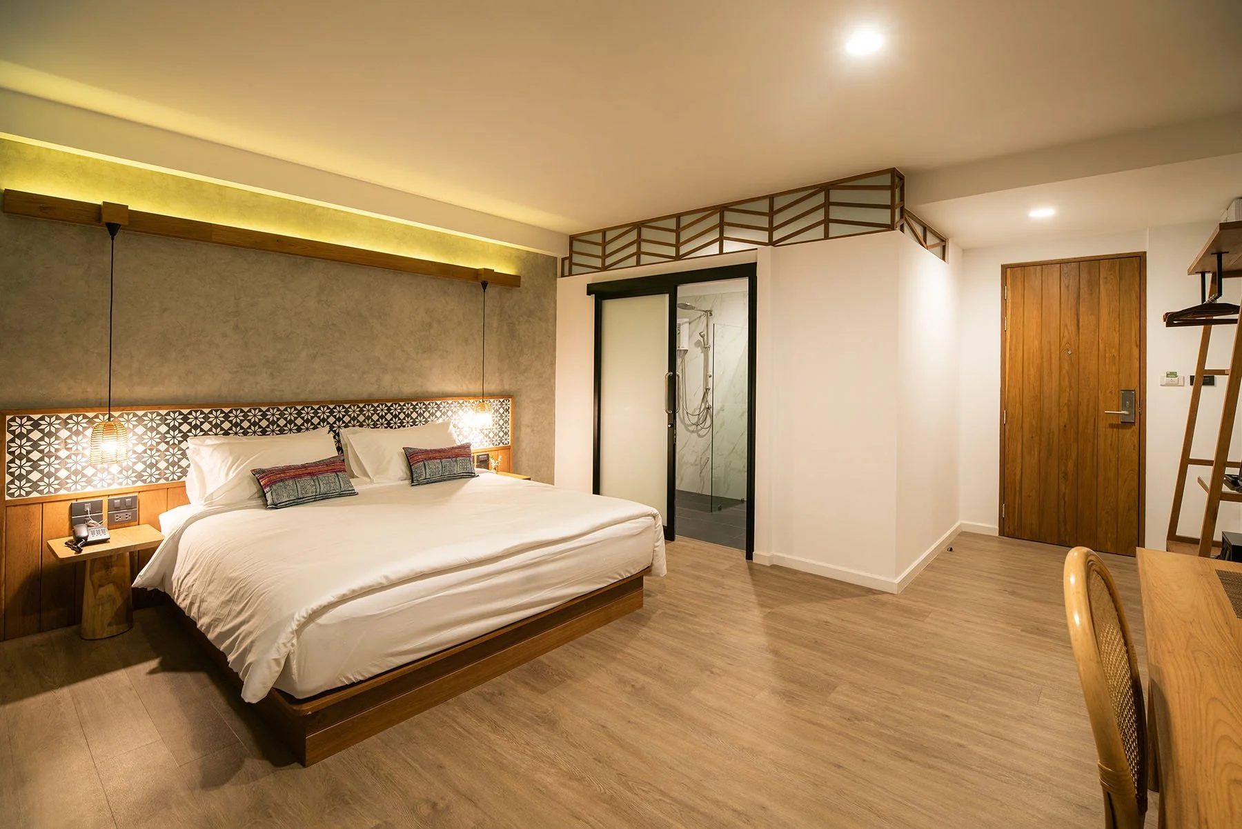 A modern hotel room with a large bed, decorative pillows, bedside table, hanging lamps, an open shower with glass door, wooden accents, and minimal decor.
