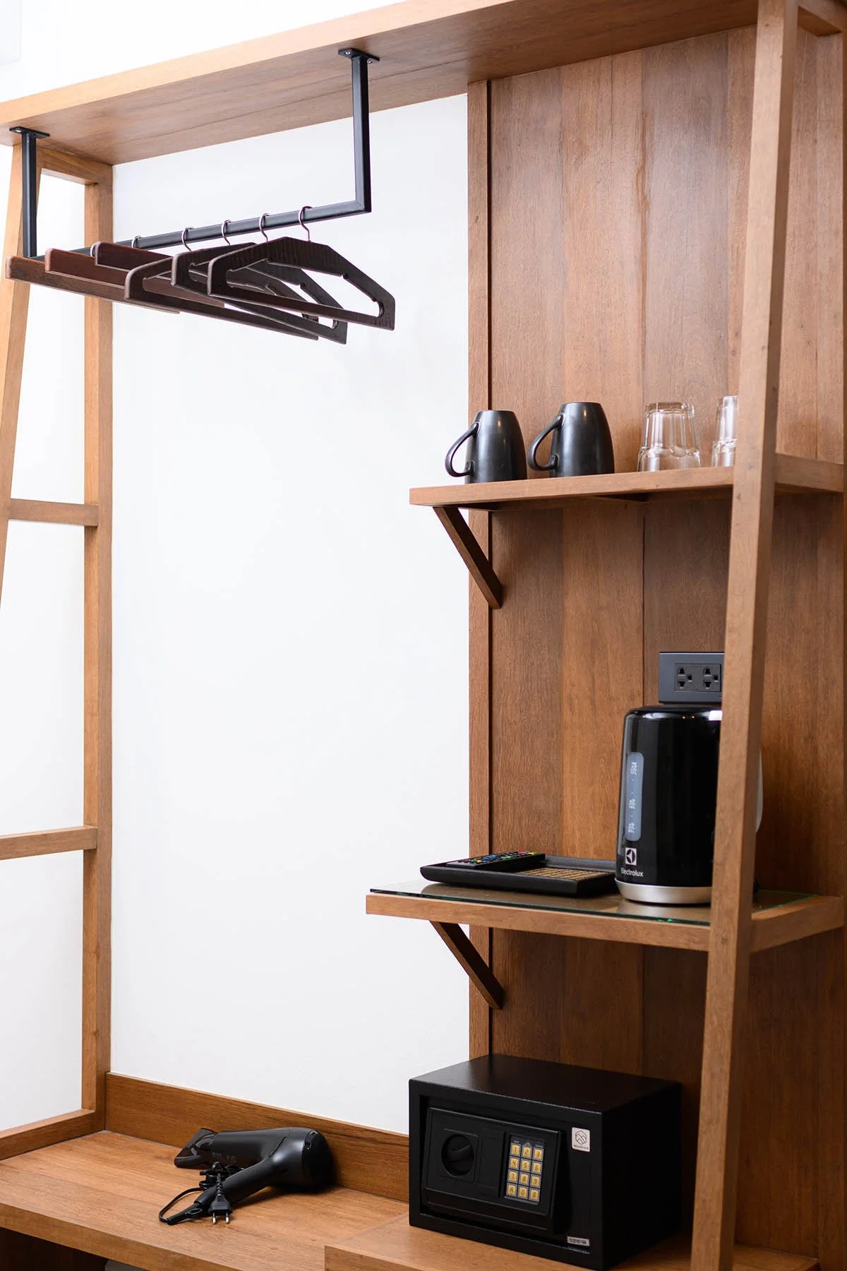 Wooden closet with hangers, electric kettle, glasses, remote, safe, and a hairbrush.