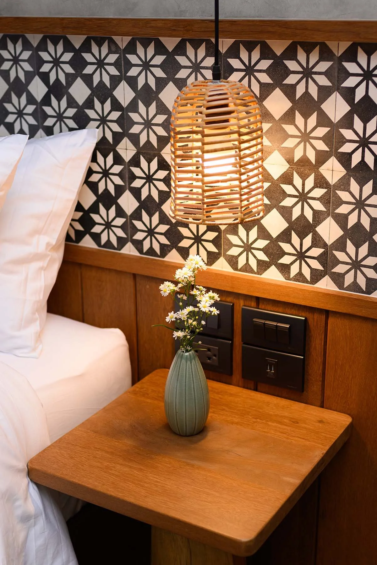 A cozy bedroom corner with a wooden nightstand holding a green vase with white flowers, a rattan pendant lamp above, and a patterned black and white tile headboard background.